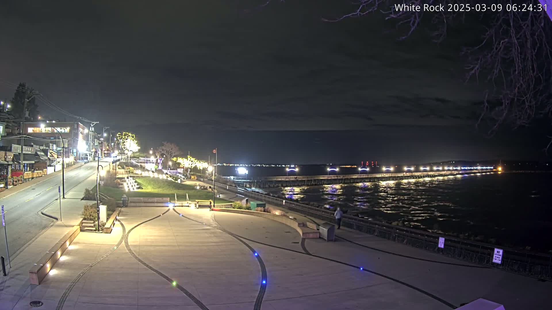 A nighttime view of a waterfront plaza with a long pier extending into a dark, choppy sea under a cloudy sky.