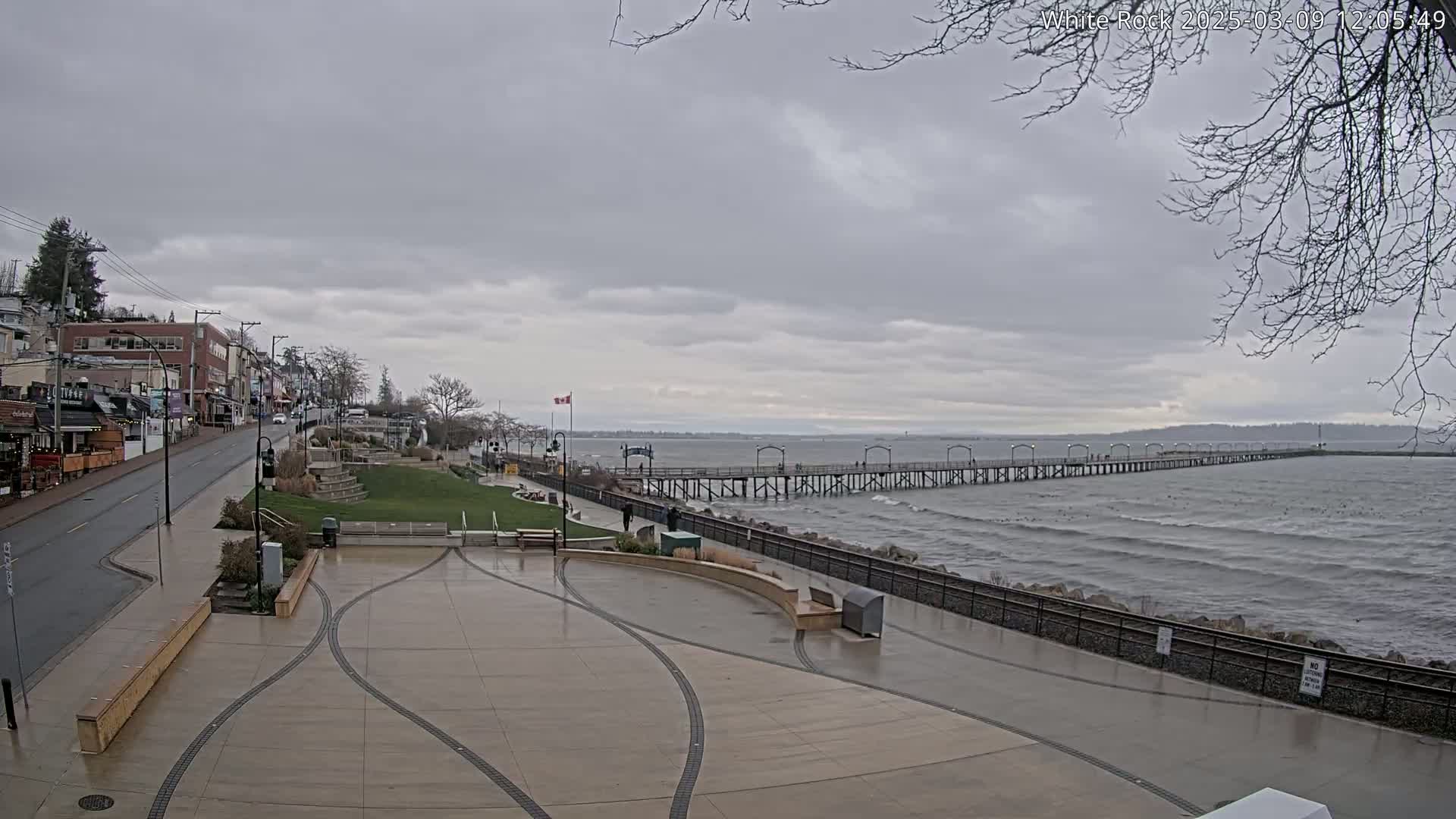 A long pier extends into a choppy sea under a cloudy sky, with a paved waterfront area and a street lined with buildings in the foreground.
