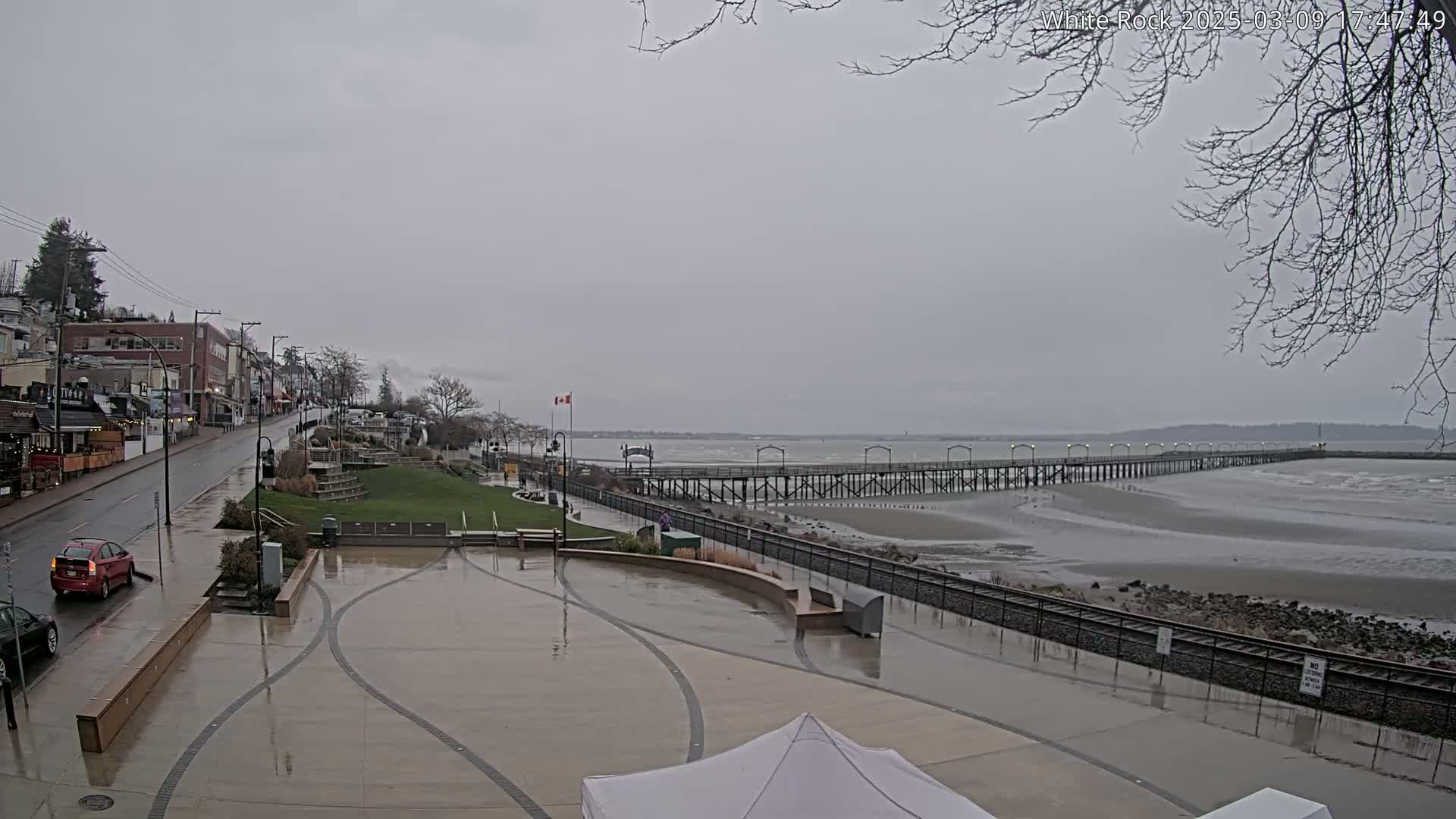 A long pier extends into a calm, grey sea under an overcast sky, adjacent to a wet paved plaza and a street lined with buildings.