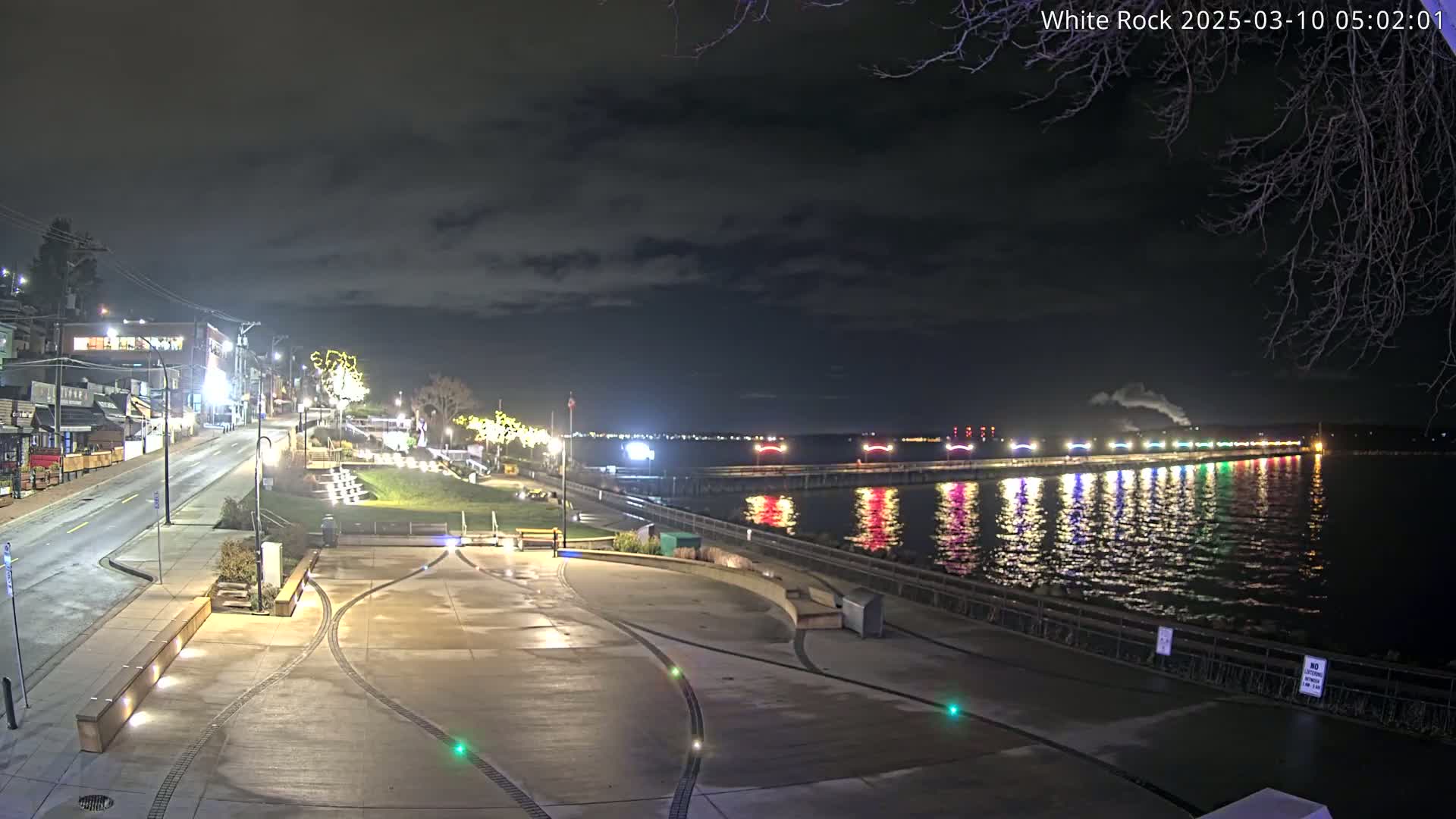A nighttime view of a waterfront plaza with a brightly lit pier extending into the calm water under a cloudy sky.