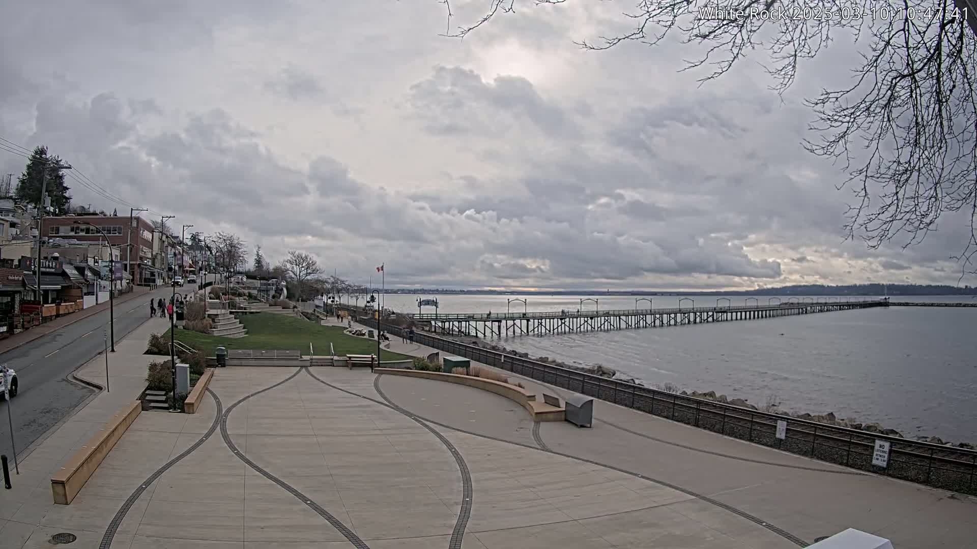 A long pier extends into a calm body of water under a cloudy sky, with a paved waterfront area and buildings visible in the background.