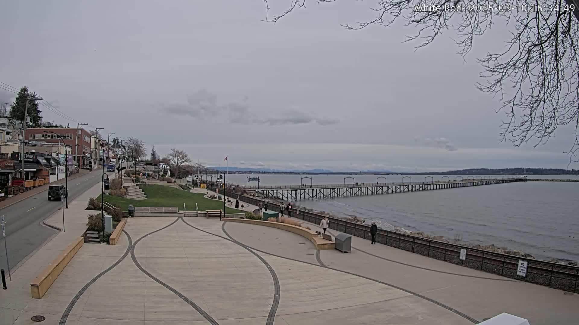 A long pier extends into a calm, grey-sky day over a body of water, with a paved waterfront area and buildings visible in the background.