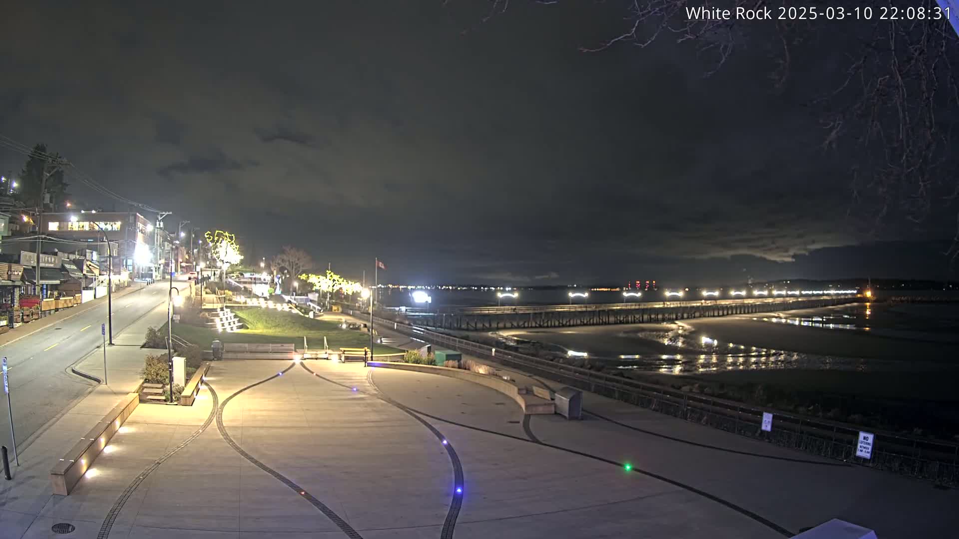 A nighttime view of a paved waterfront area with benches and lights, overlooking a long pier extending into a dark, calm body of water under a cloudy sky.