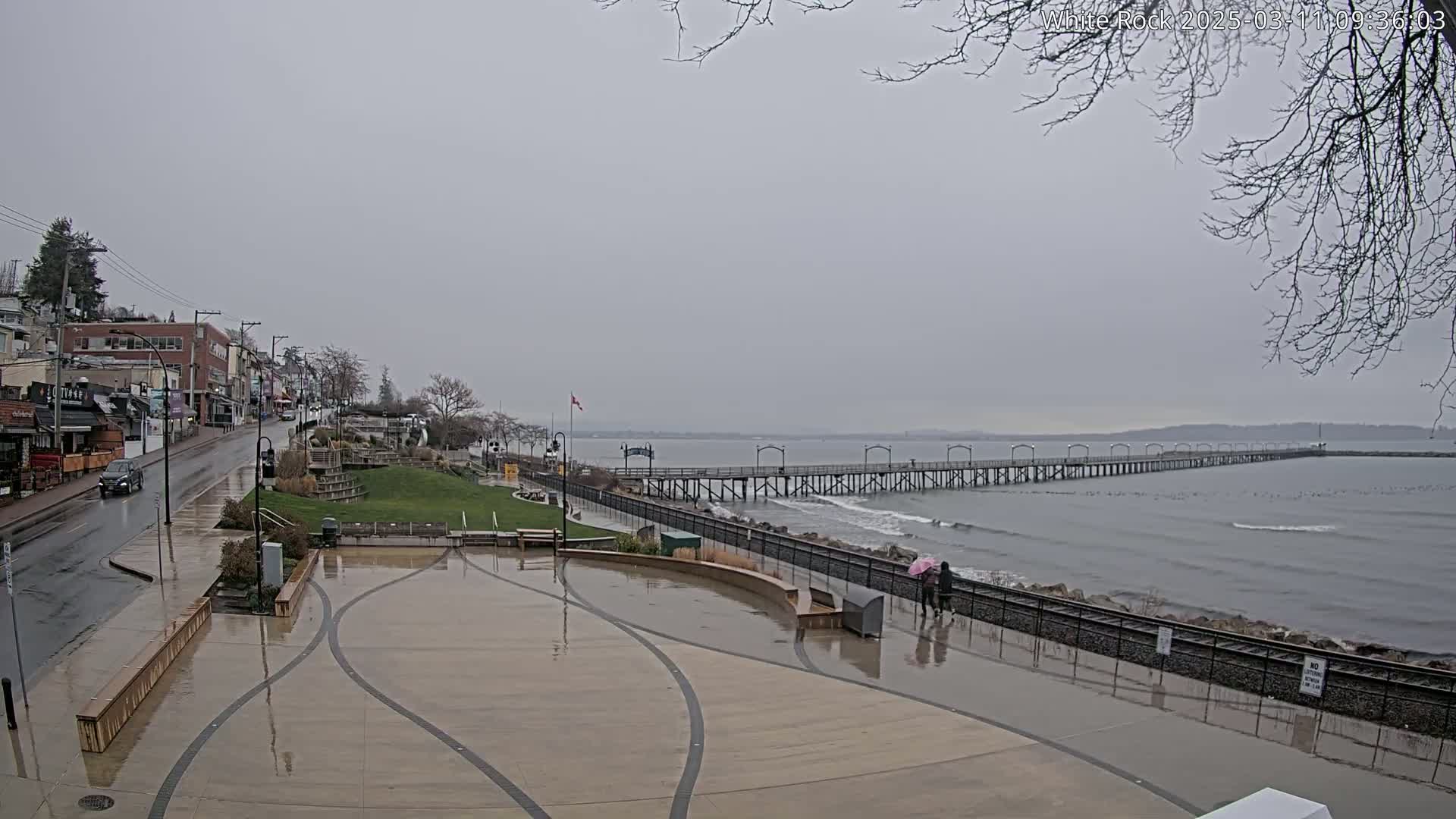A wet, paved plaza overlooks a long pier extending into a gray, choppy ocean under an overcast sky.