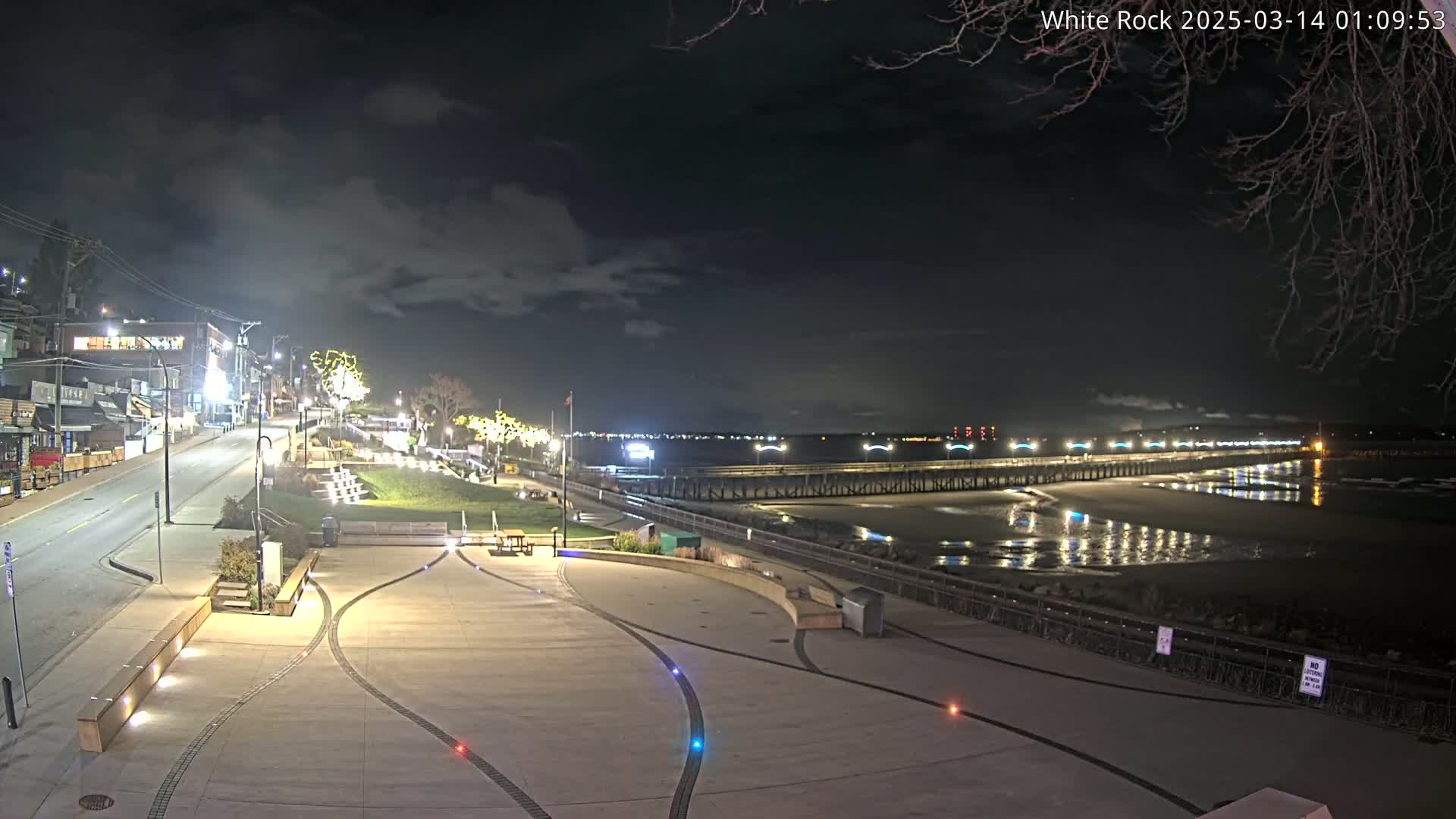 A nighttime view of a paved waterfront plaza, featuring a long pier extending into calm water under a partly cloudy sky.