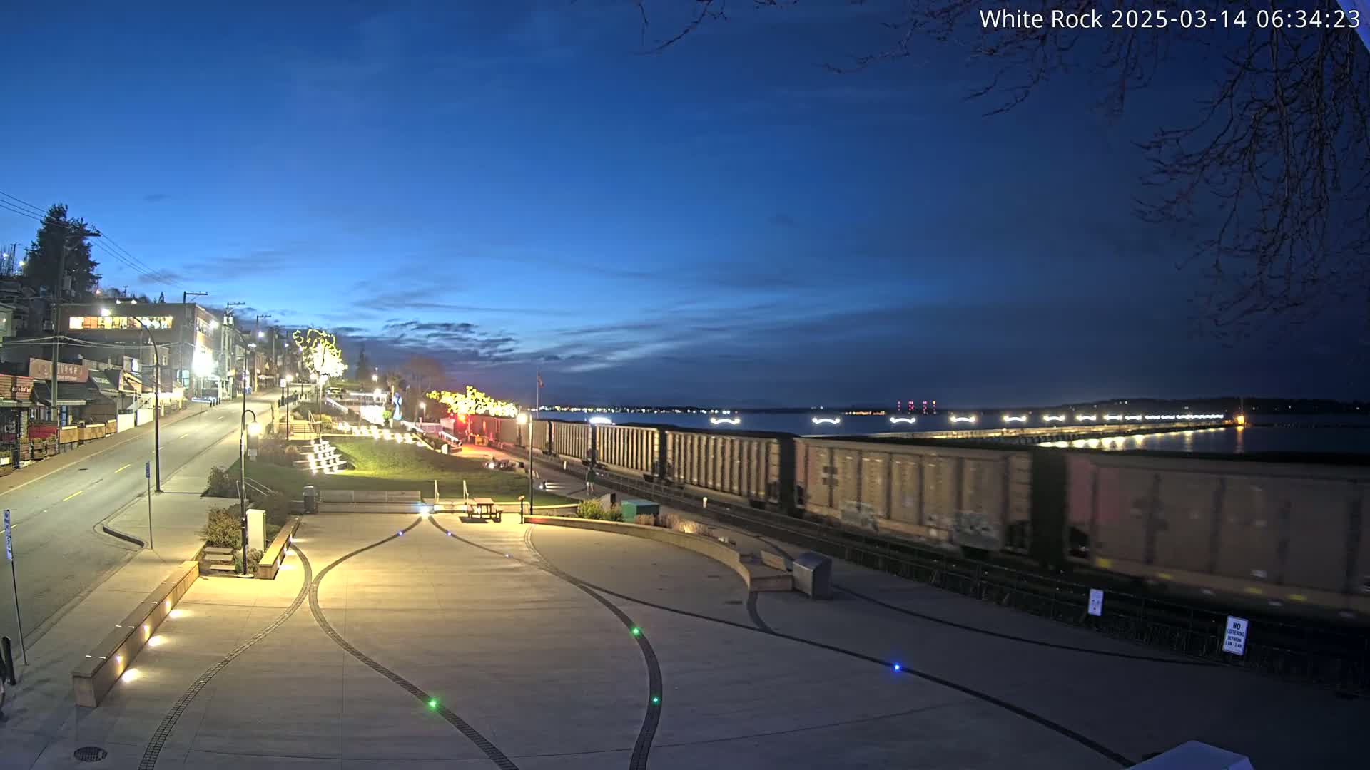 A long freight train passes by a waterfront plaza at twilight under a clear, dark blue sky.