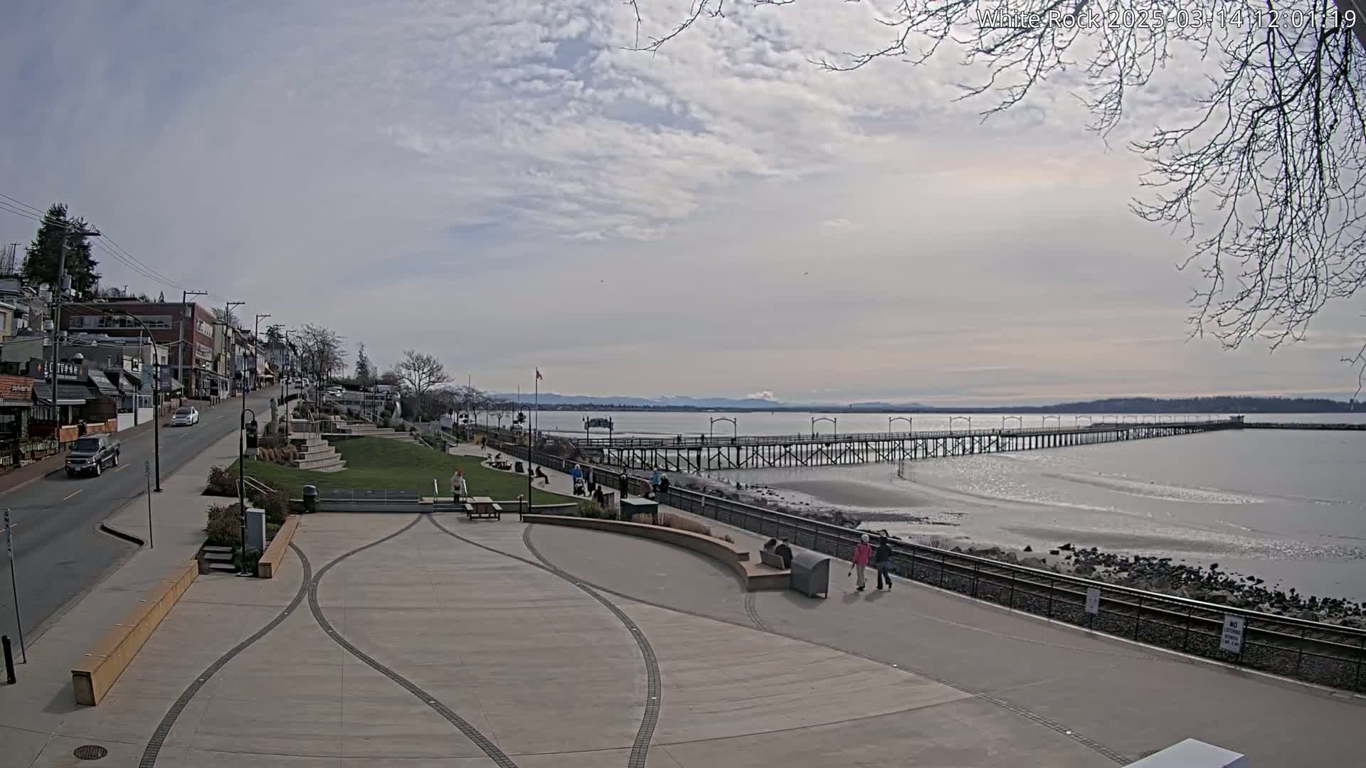 A long pier extends into a calm, gray body of water under a partly cloudy sky, with a paved waterfront area and a street lined with buildings in the foreground.