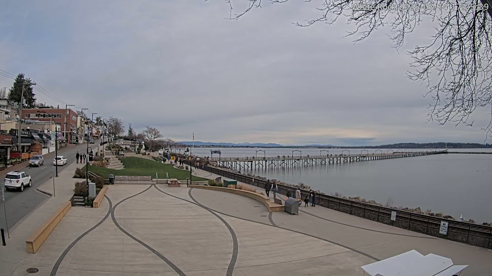 A long pier extends into a calm, gray body of water under an overcast sky, with a paved waterfront area and buildings visible in the foreground.