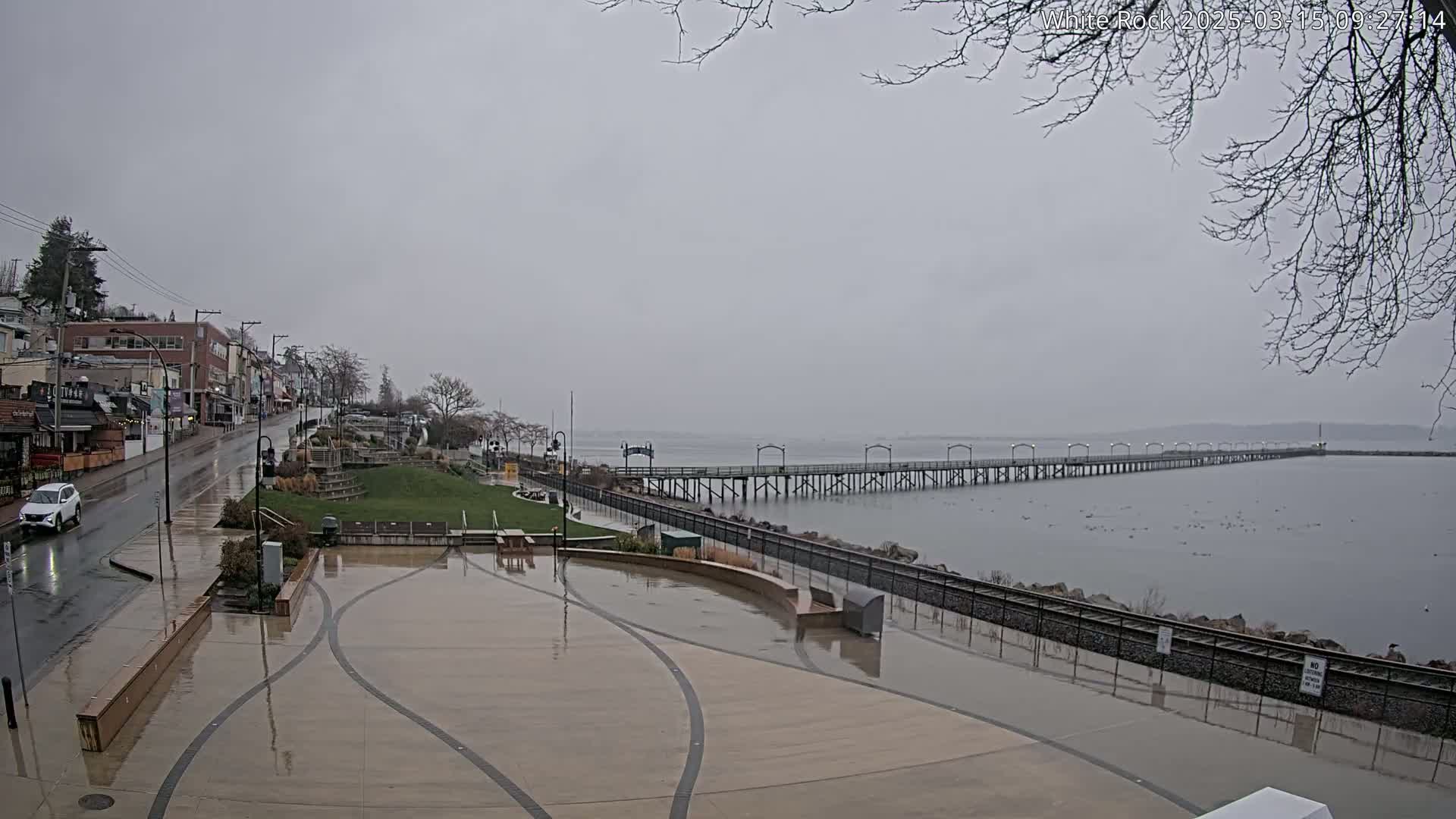 A long pier extends over calm water under a gray, overcast sky, next to a wet paved area and buildings, with a car on a rain-slicked street in the foreground.