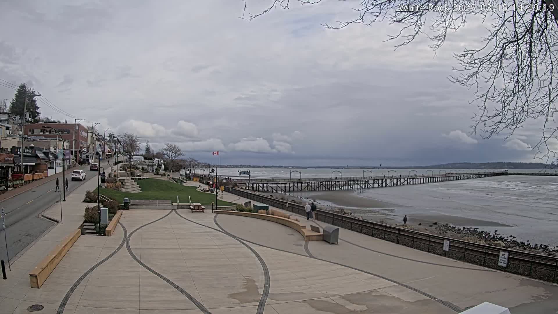 A long pier extends into a choppy sea under a cloudy sky, with a paved waterfront area and buildings visible in the foreground.