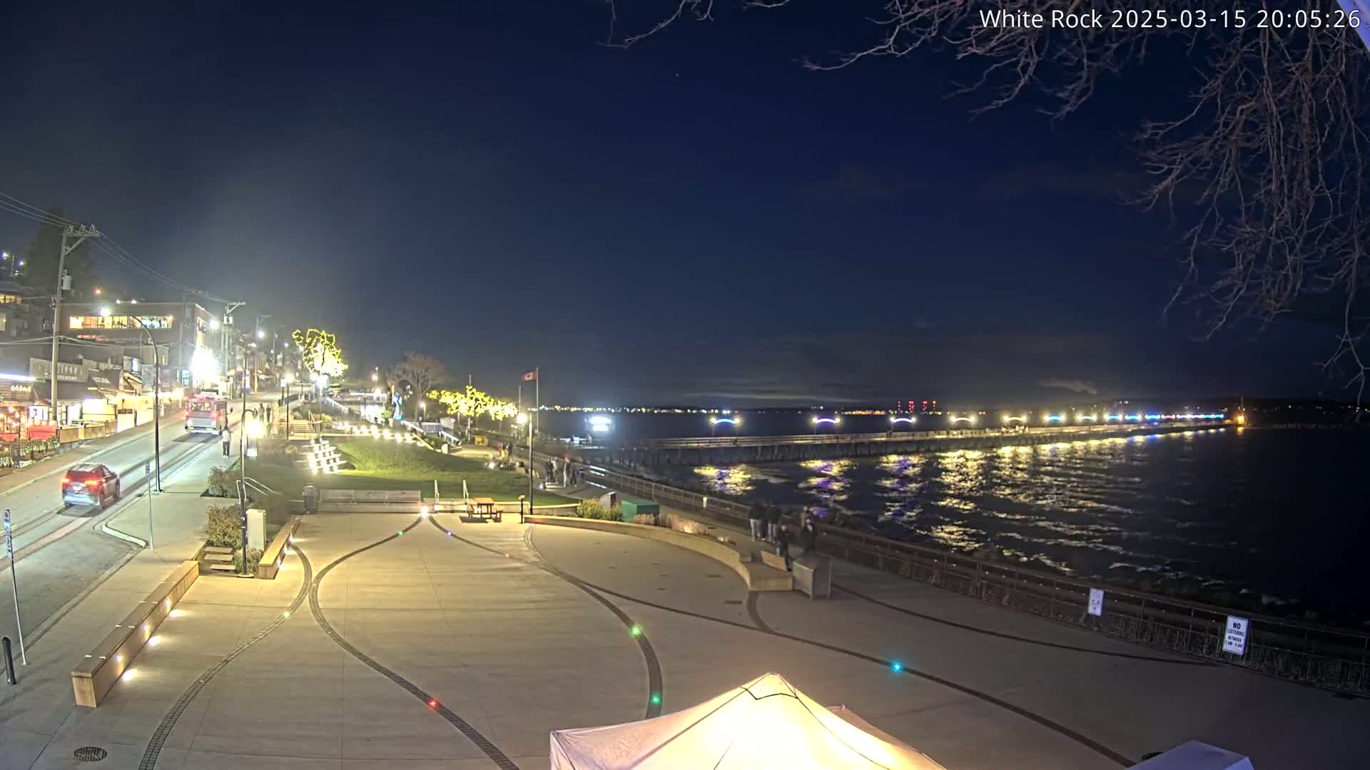 A nighttime view of a waterfront plaza with a long pier extending into a dark, somewhat choppy body of water under a clear, dark sky.