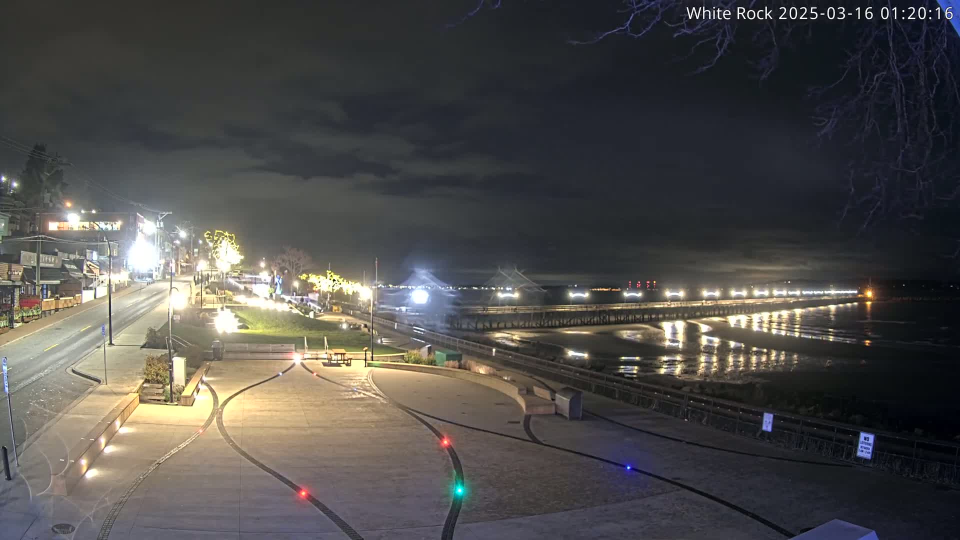 A nighttime view of a paved waterfront area with a long pier extending into a calm, dark ocean under a partly cloudy sky.