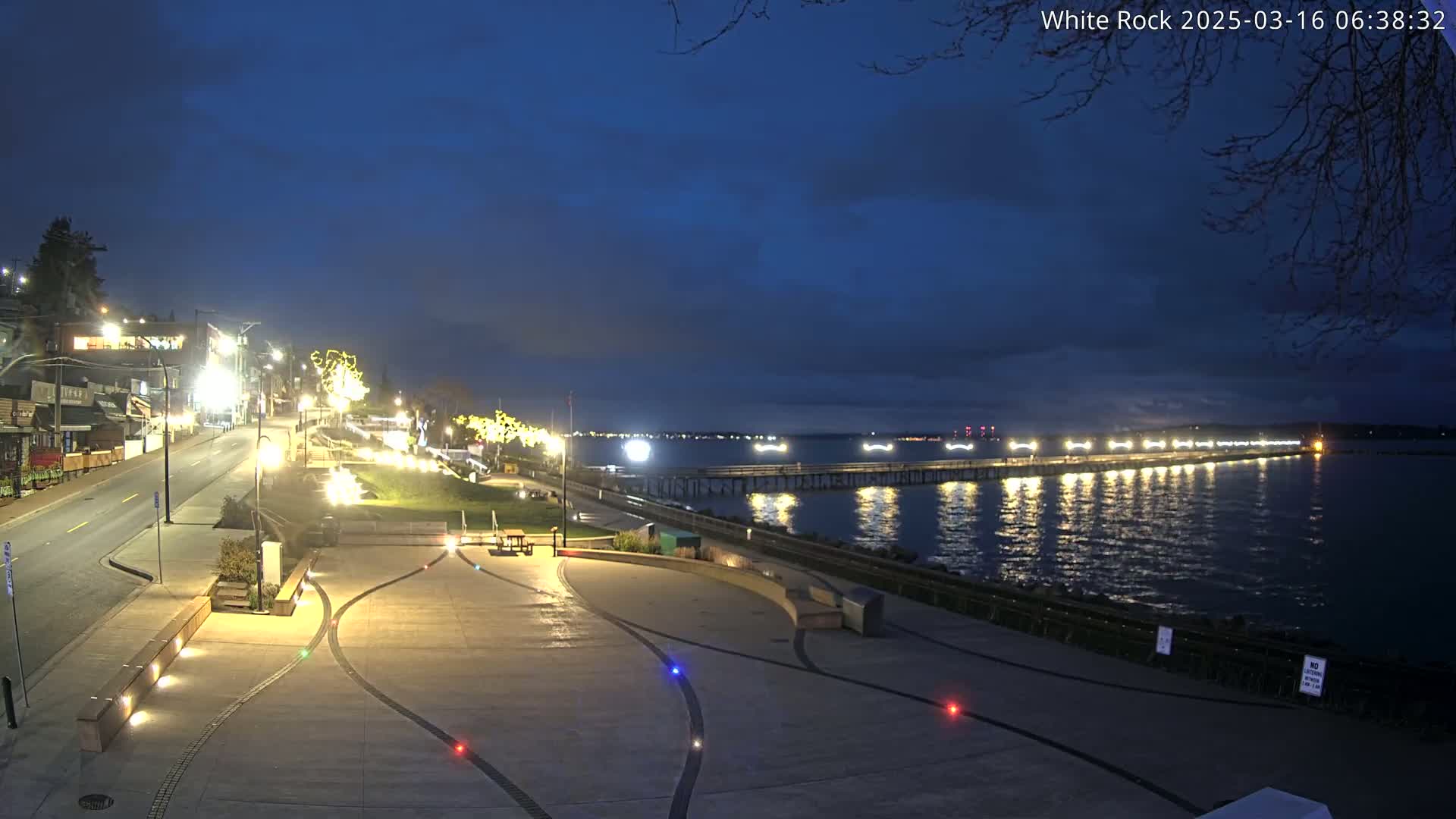 A brightly lit waterfront plaza at night, overlooking a pier extending into a calm, dark body of water under a partly cloudy sky.