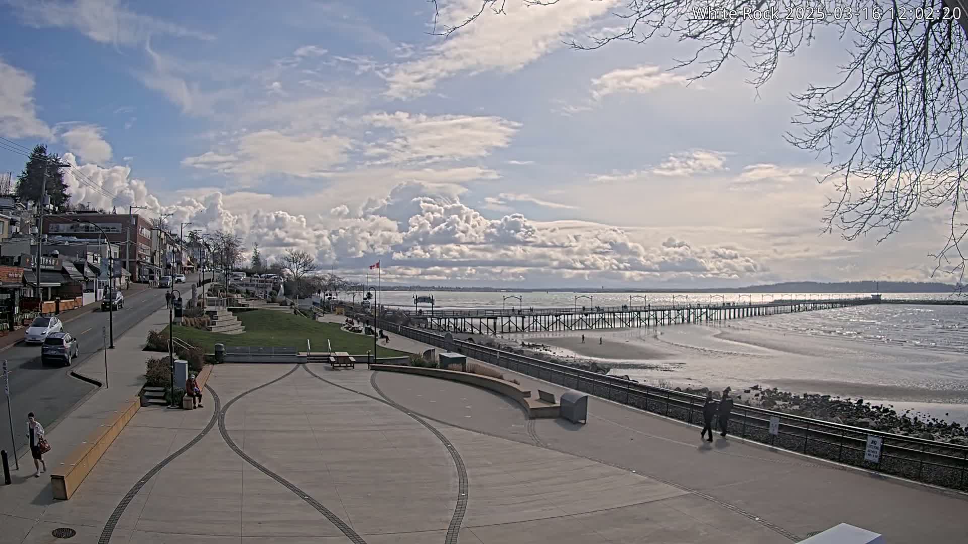 A long pier extends into a calm, shallow ocean under a partly cloudy sky, with a paved waterfront area and buildings in the foreground.