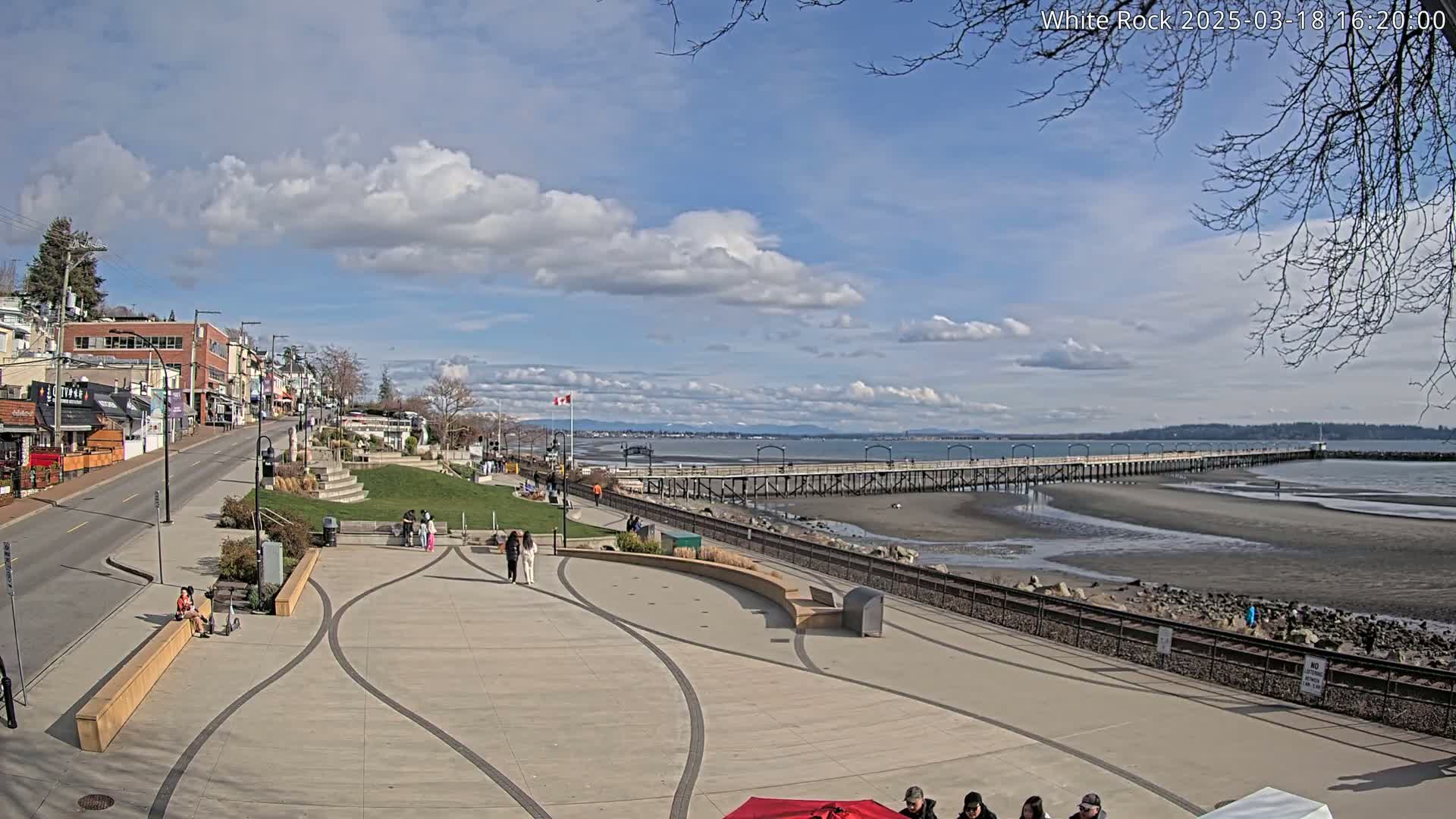 A paved waterfront plaza with a long pier extending into a partly exposed sandy bay under a partly cloudy sky.