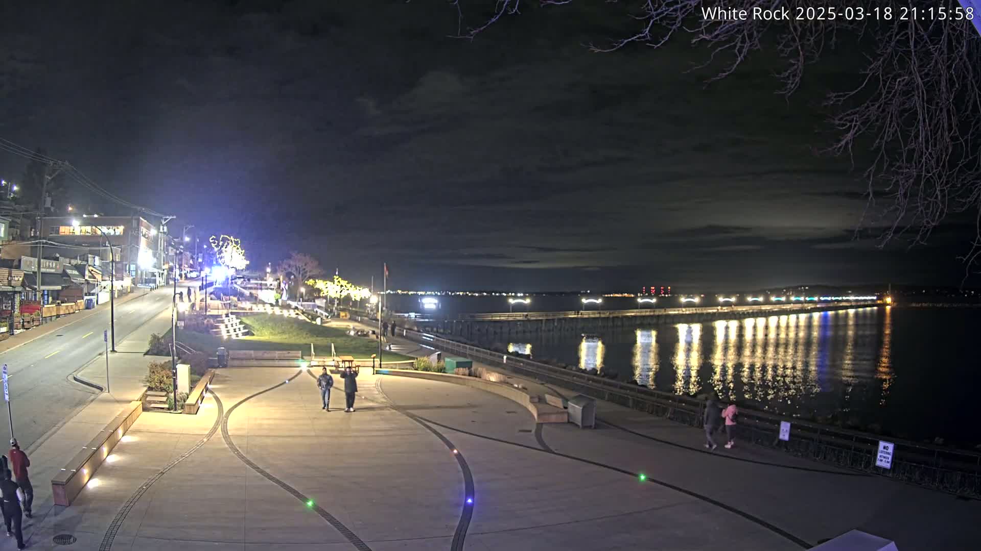 A nighttime view of a waterfront plaza with a few people walking, overlooking a long, illuminated pier extending into calm water under a dark, cloudy sky.