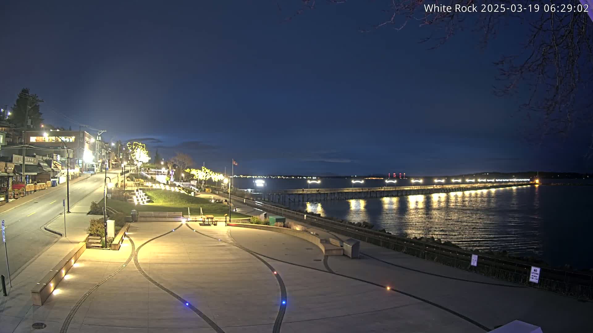 A nighttime view of a waterfront town with a long pier extending into a calm, dark water, under a clear, dark blue sky.