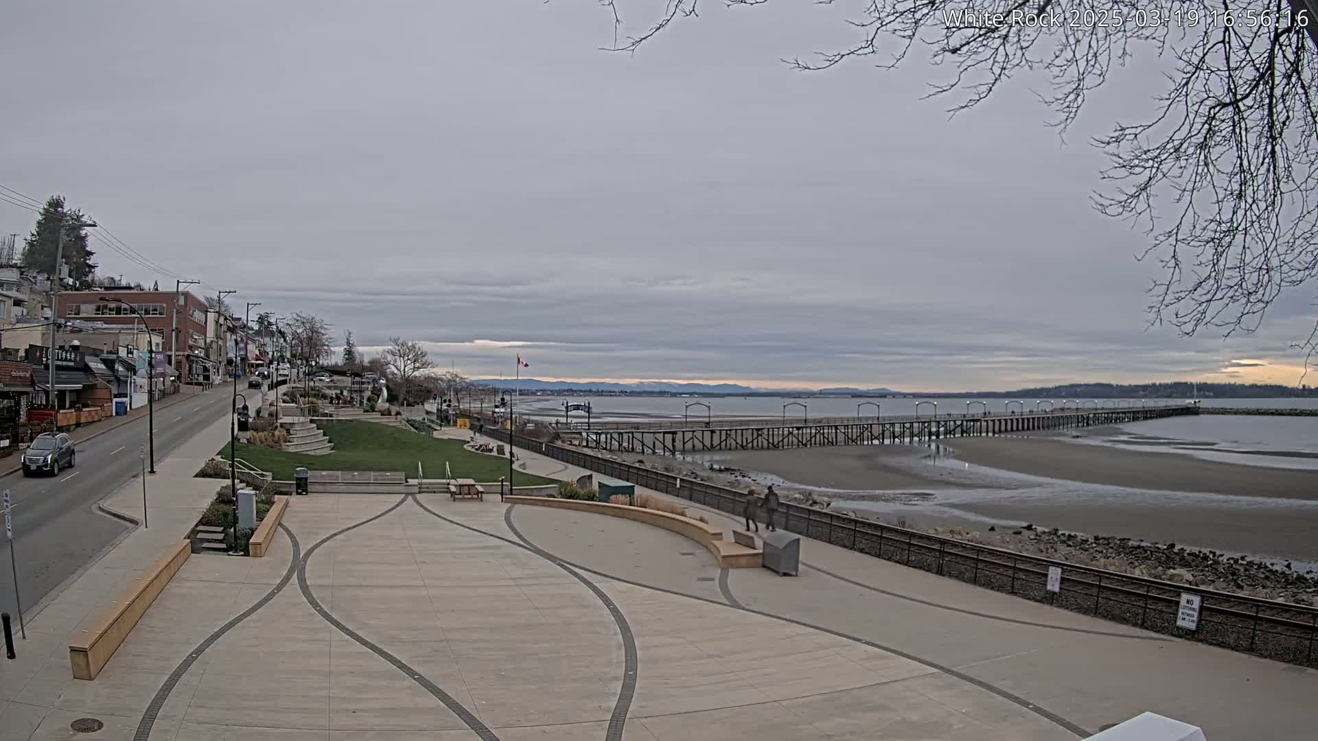 A paved plaza overlooks a long pier extending into a calm, overcast ocean, with a town visible in the background.