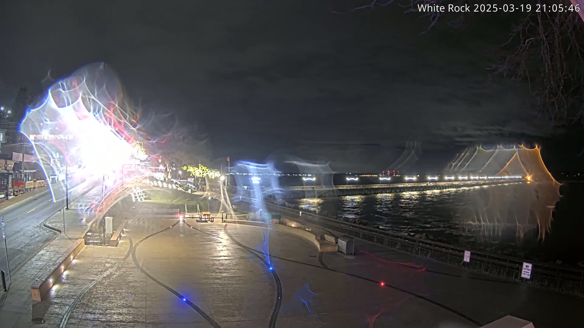 A nighttime view of a waterfront plaza with light trails, a body of water, and a pier with lights in the background under a partly cloudy sky.