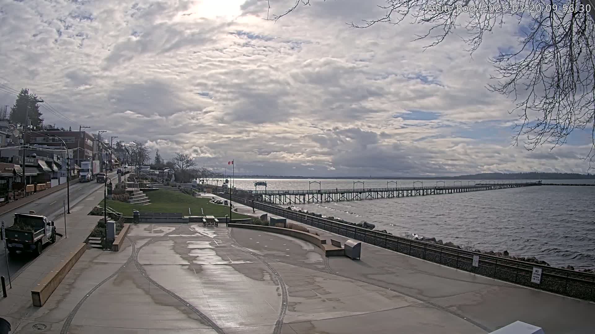A mostly cloudy day shows a seaside town with a long pier extending into the water, a paved waterfront area, and buildings along a street.