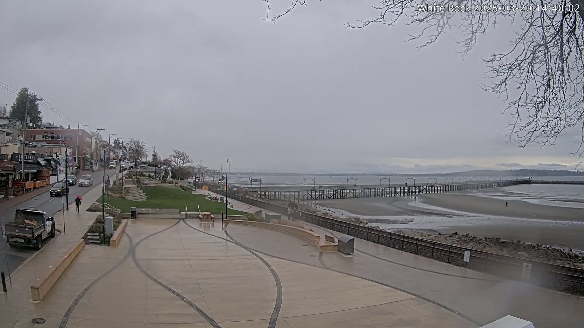 A wet, gray day reveals a seaside town with a long pier extending into a calm, exposed bay, viewed from a paved plaza overlooking the water.