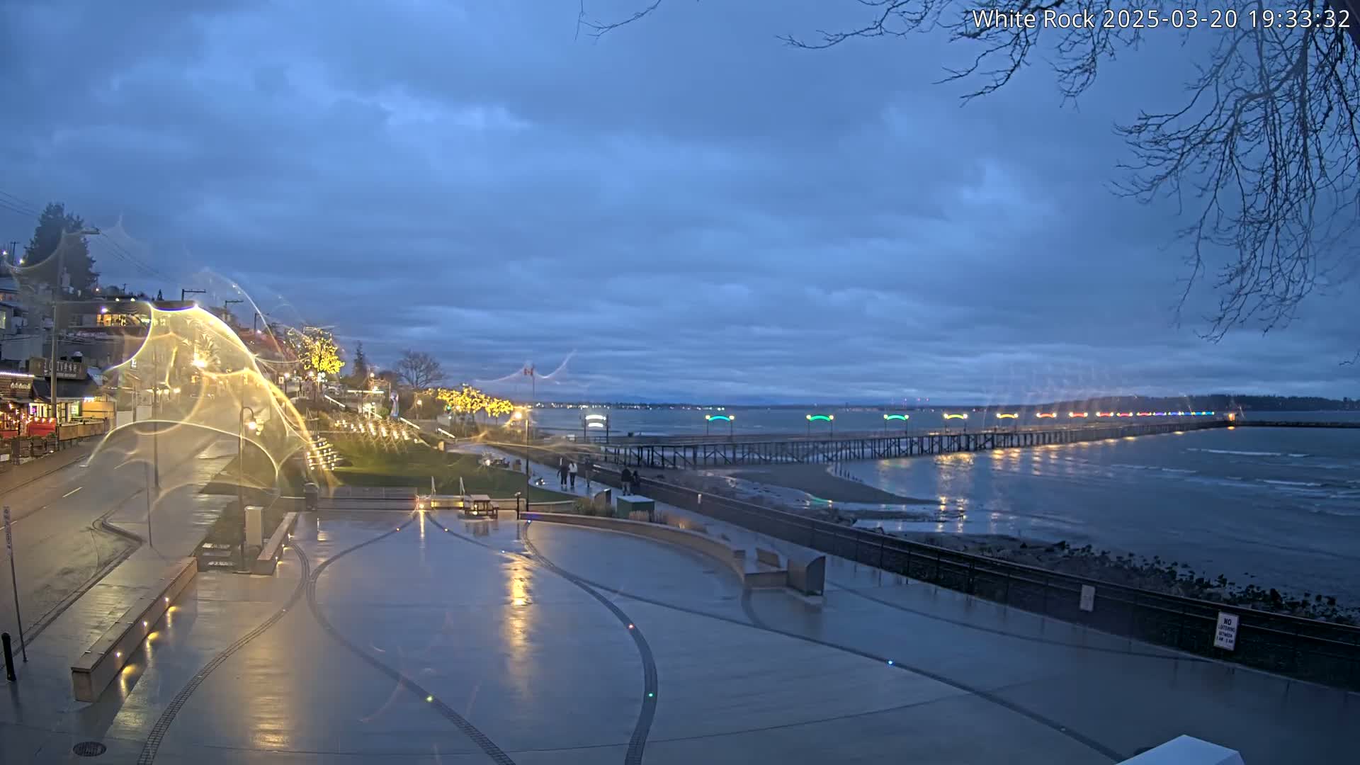 A wet, paved plaza with lights is shown at dusk, overlooking a long pier with colorful lights illuminating it at the edge of a calm, dark-blue body of water under a cloudy sky.