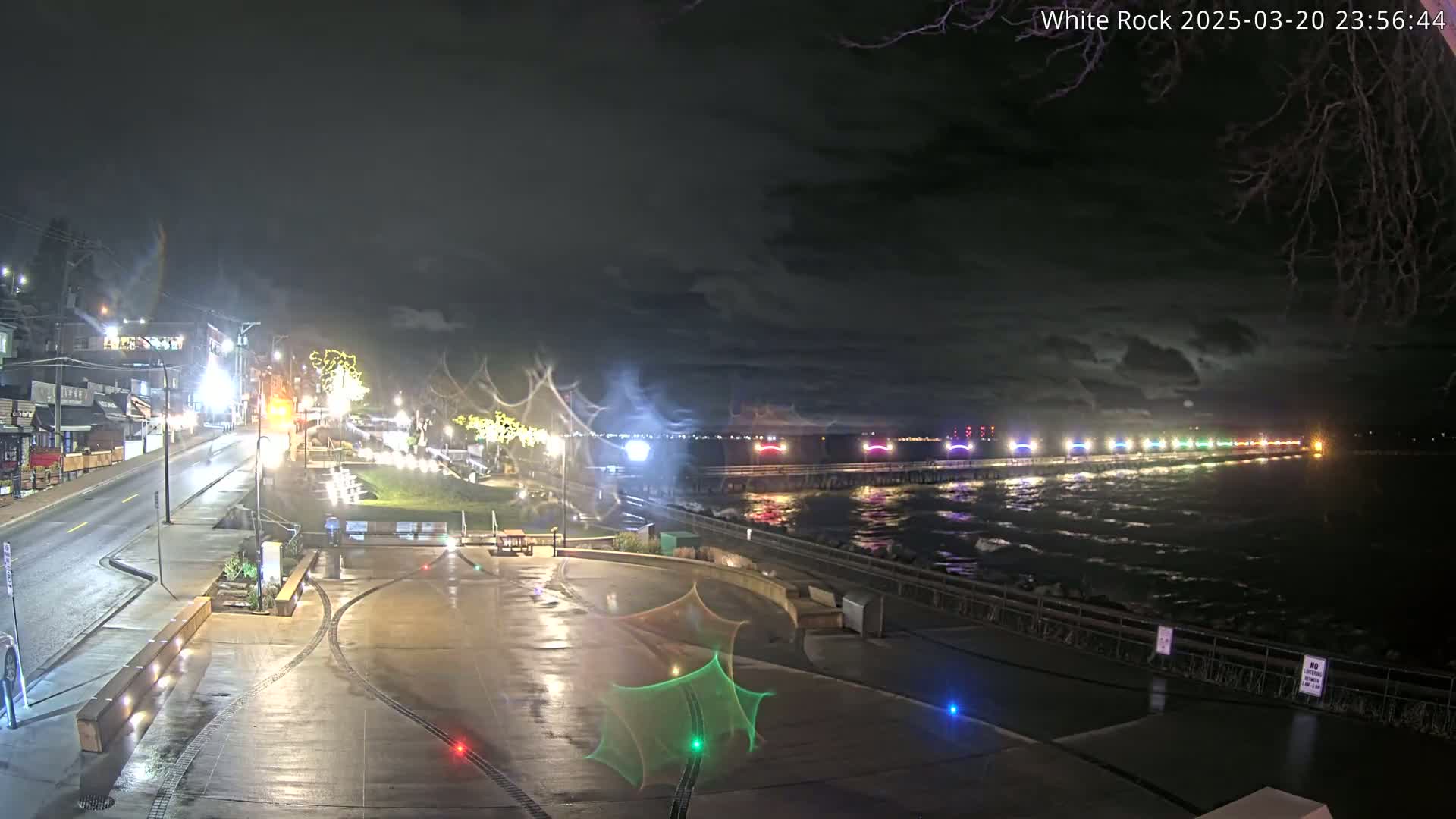 A nighttime view of a wet, paved waterfront area with a pier lit with colorful lights extending into a dark, choppy body of water under a cloudy sky.