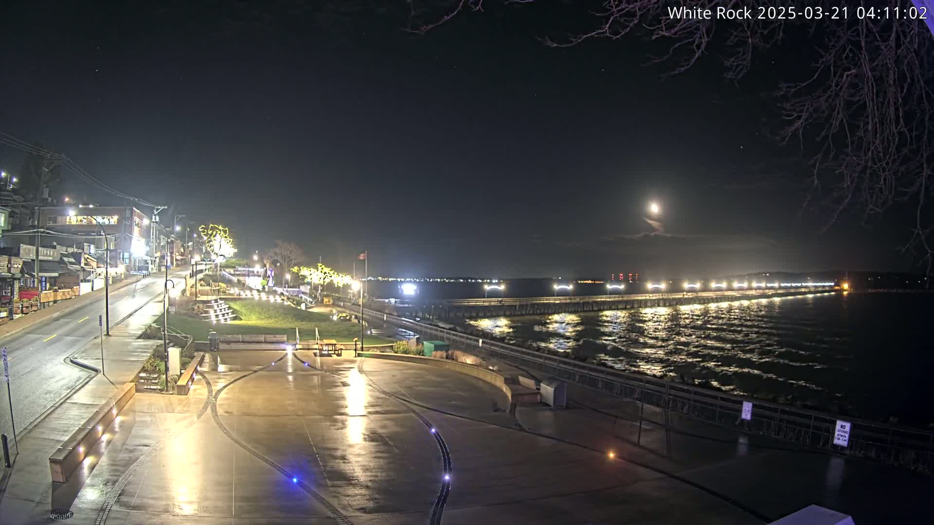 A nighttime view of a waterfront town, featuring a long pier with lights extending into a dark, wavy ocean under a moonlit sky.
