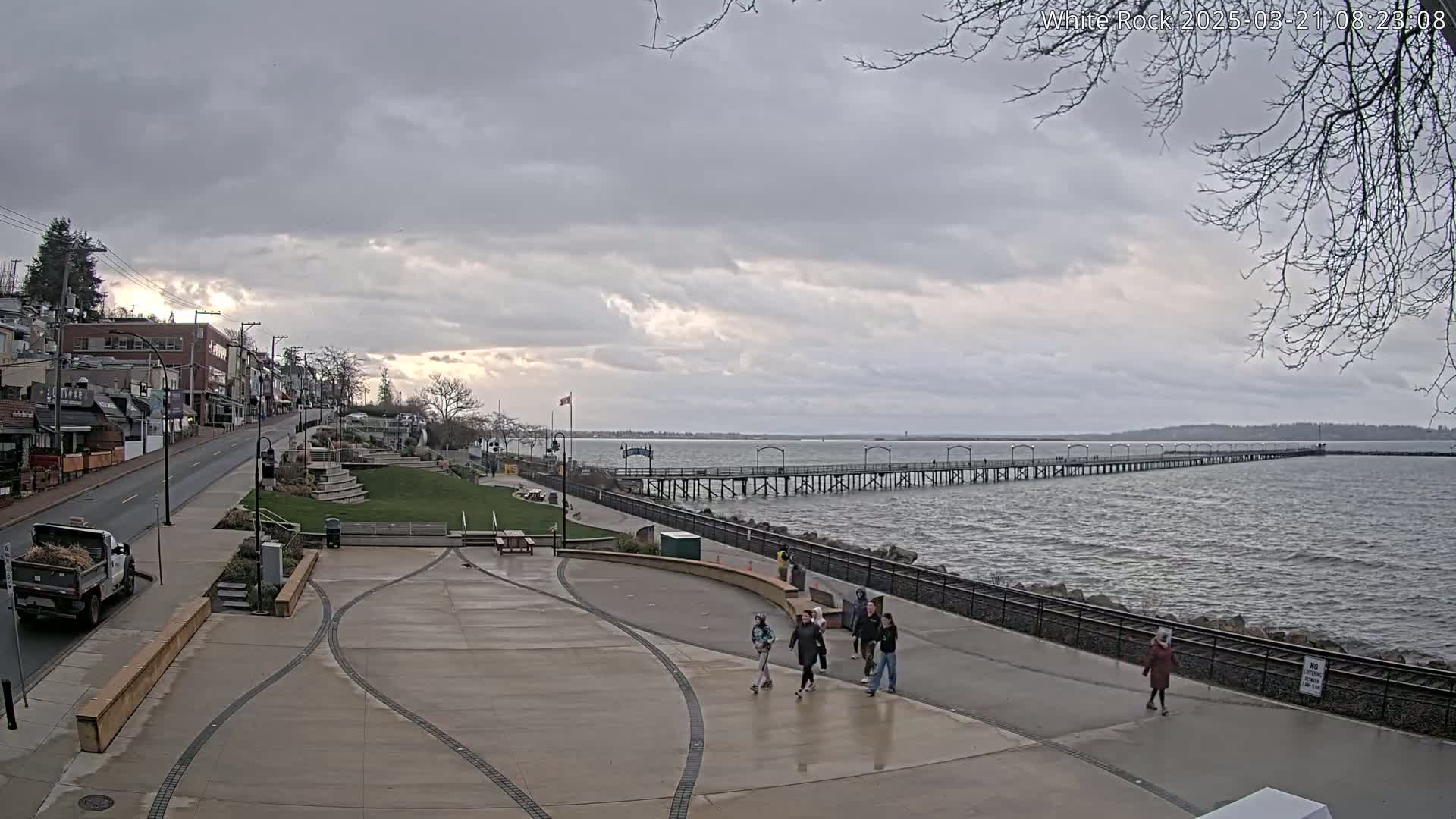 A long pier extends into a gray, choppy ocean under a cloudy sky, with a paved waterfront area and a street lined with buildings visible in the foreground.