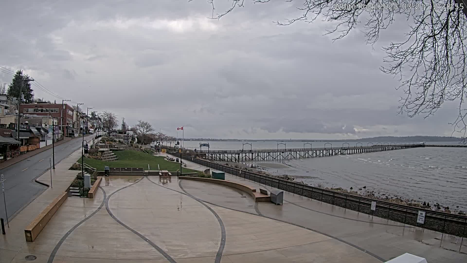 A long pier extends into a gray, choppy ocean under an overcast sky, with a paved waterfront area and buildings visible in the foreground.