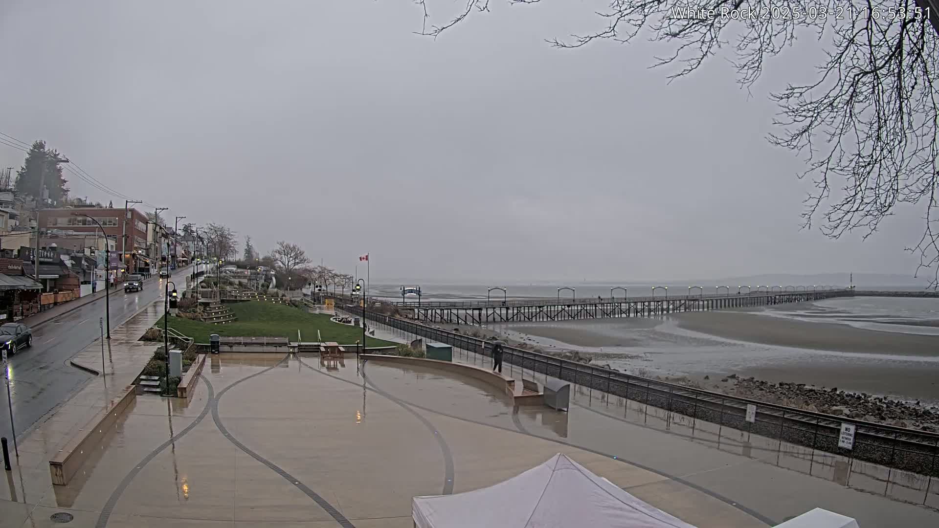 A long pier extends into a calm, gray sea under an overcast sky, with a wet town square and street in the foreground.