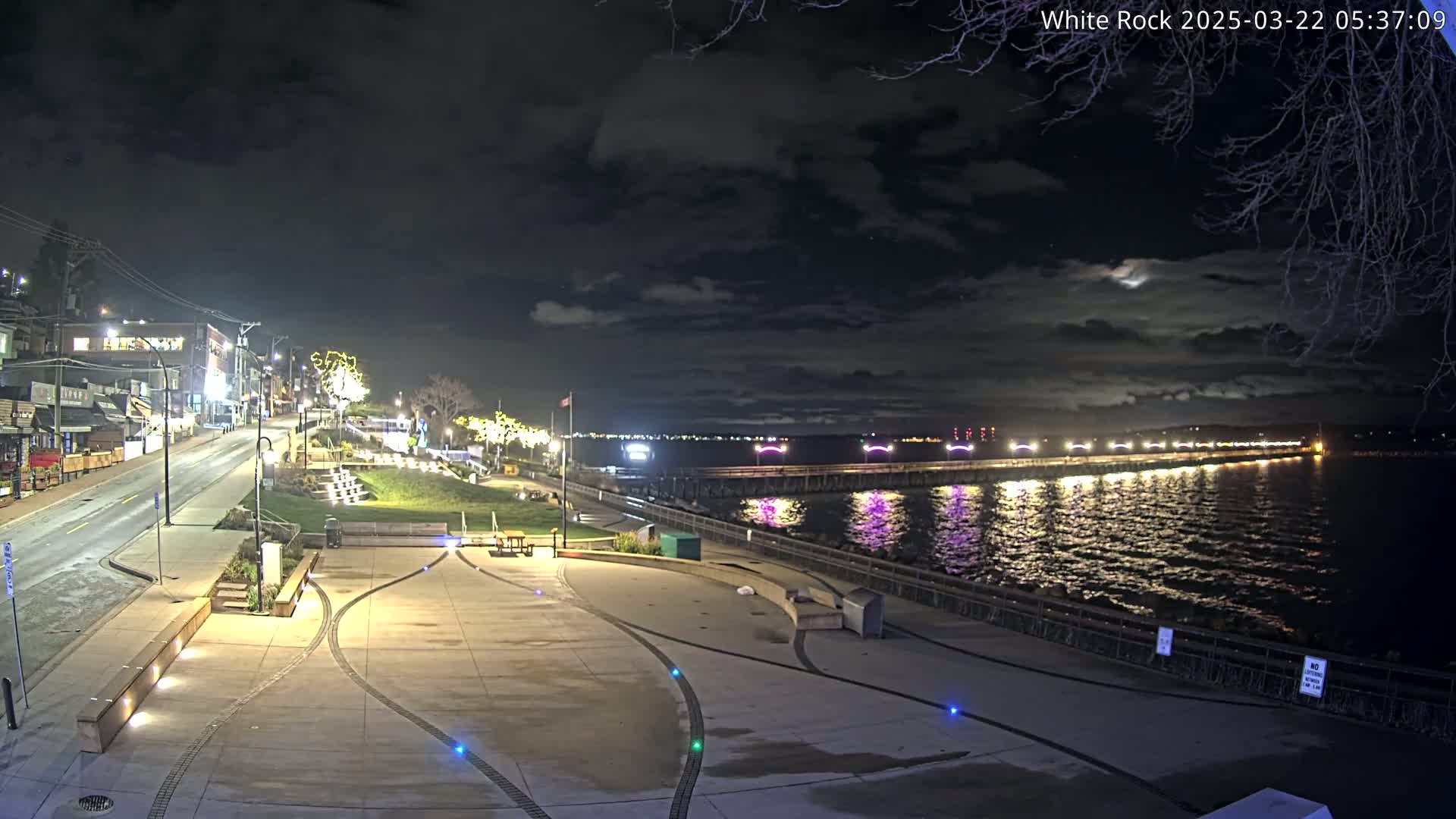 A nighttime view of a waterfront plaza with a street and buildings in the background, under a cloudy sky.