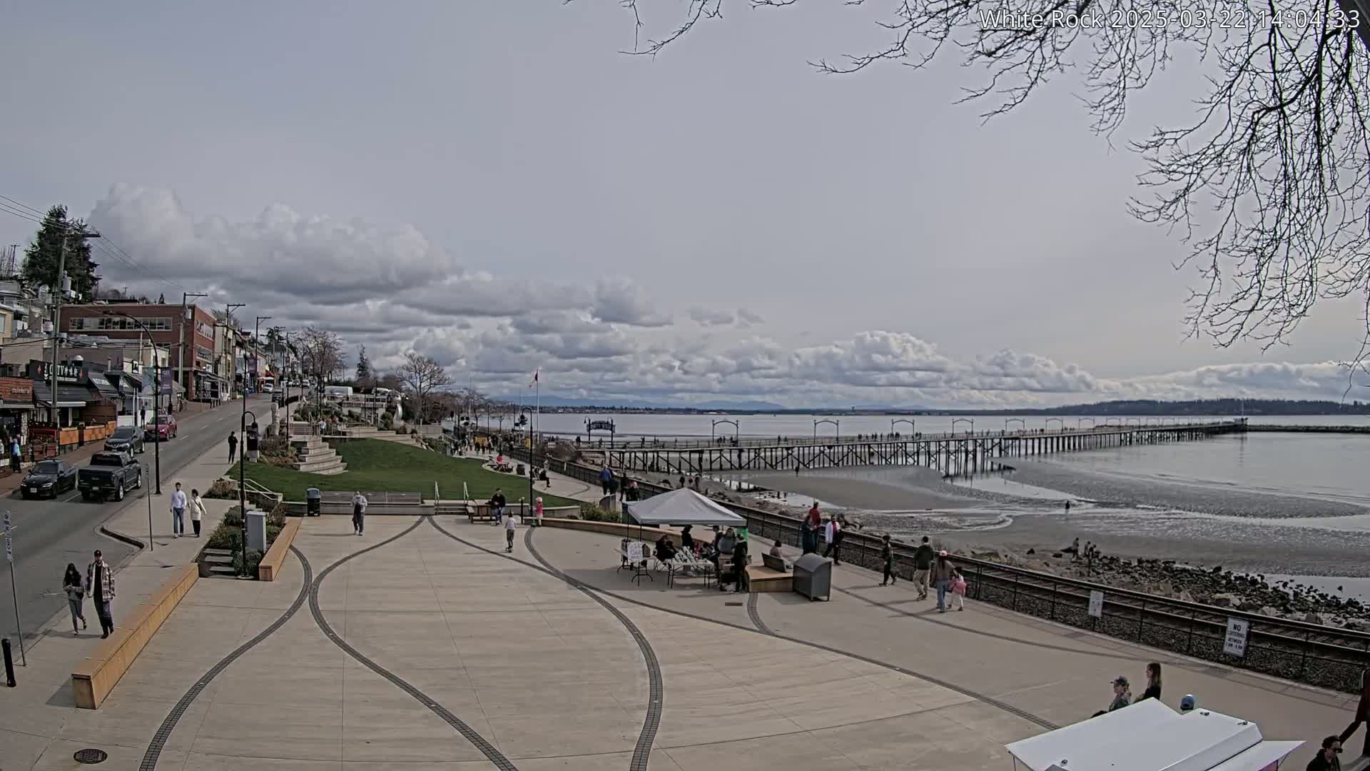 A paved waterfront area with a long pier extending into a calm bay under a partly cloudy sky, featuring people strolling along the paths and beach.