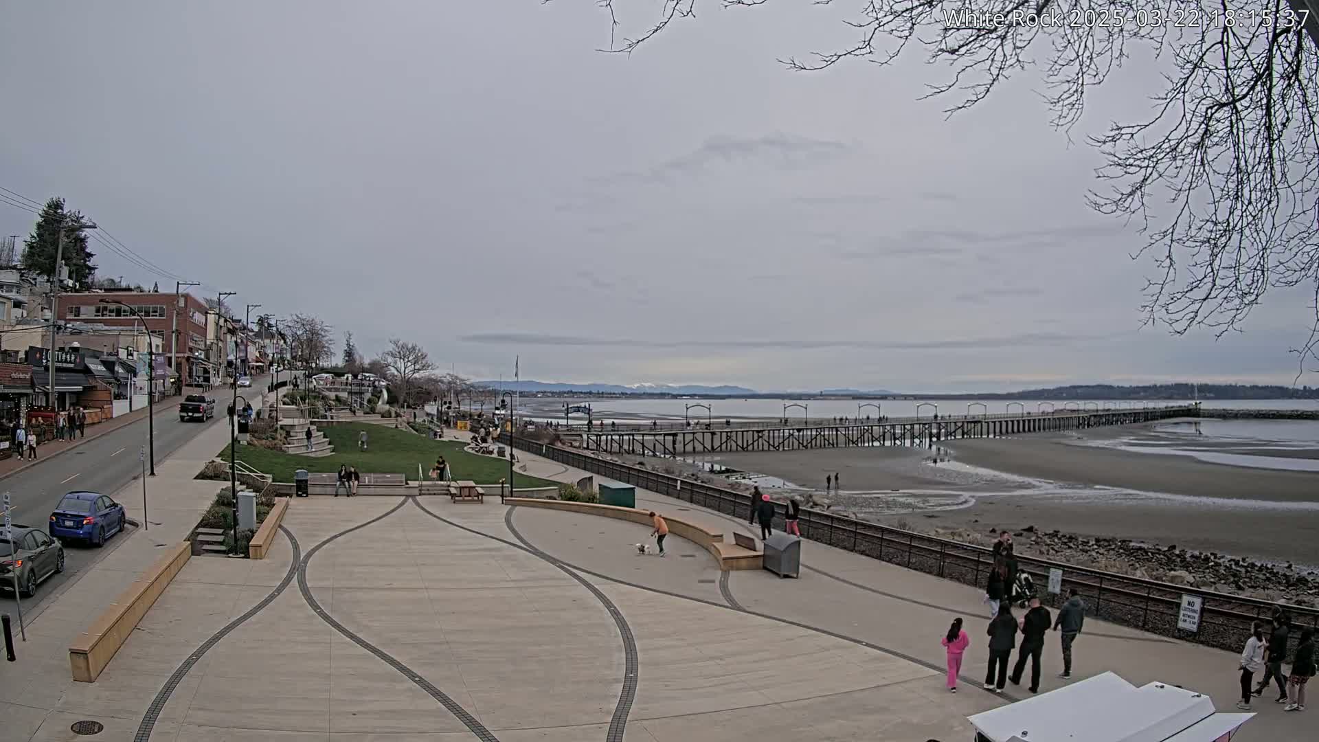 A long pier stretches across a wide, sandy beach under a cloudy sky, with a paved waterfront promenade and shops visible in the background.