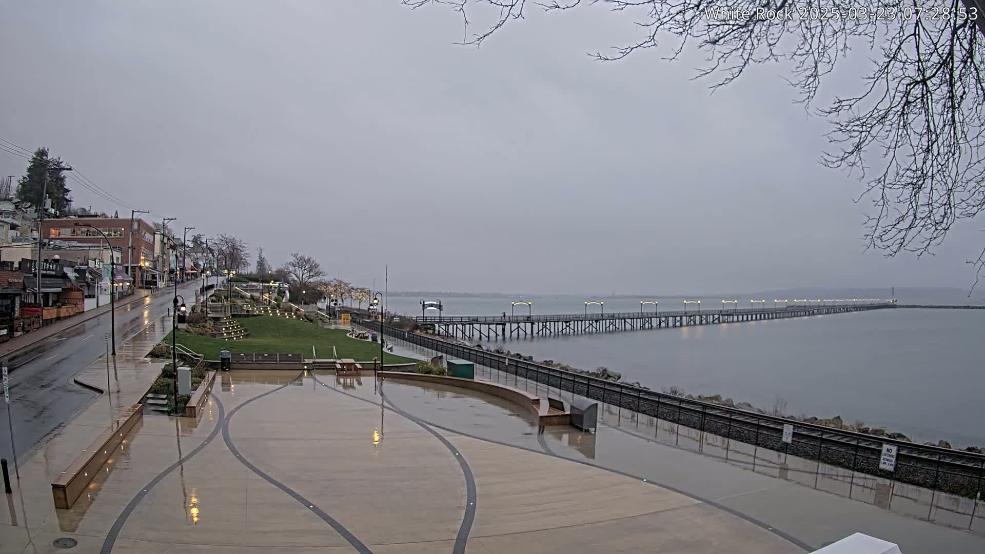 A long pier extends into a calm, gray body of water under an overcast sky, with a rain-slicked plaza and street visible in the foreground.