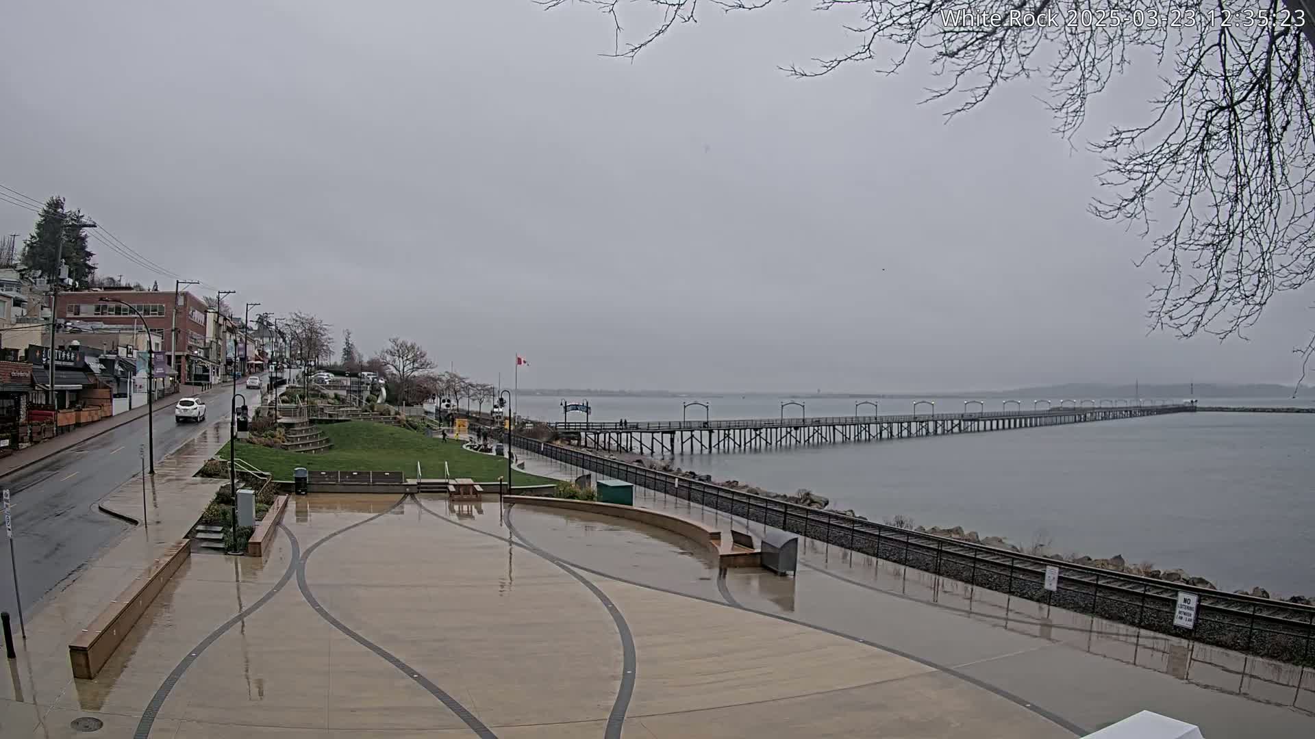 A wet plaza overlooks a long pier extending into a calm, gray body of water under an overcast sky.