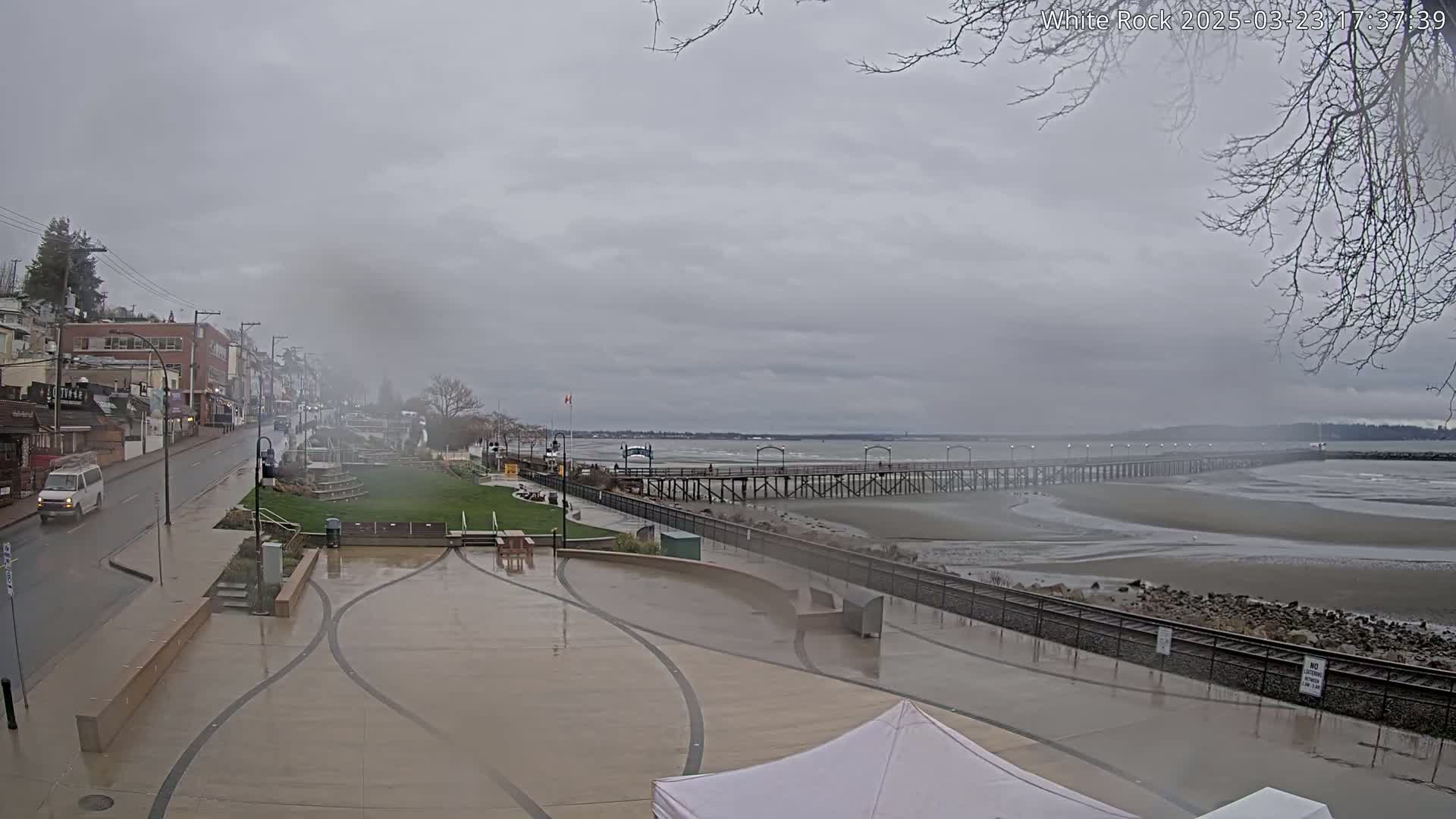A wet, overcast day reveals a town's waterfront, featuring a long pier extending into a tidal flat, and a street lined with buildings.