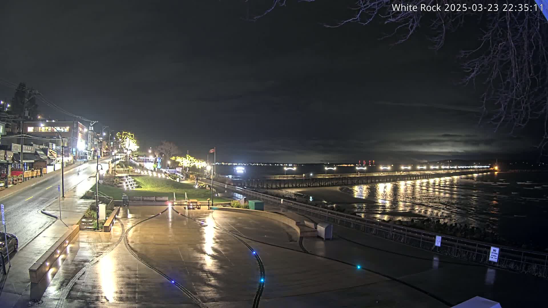 A nighttime view of a wet, paved waterfront area with a long pier extending into a dark, wavy ocean under a cloudy sky.