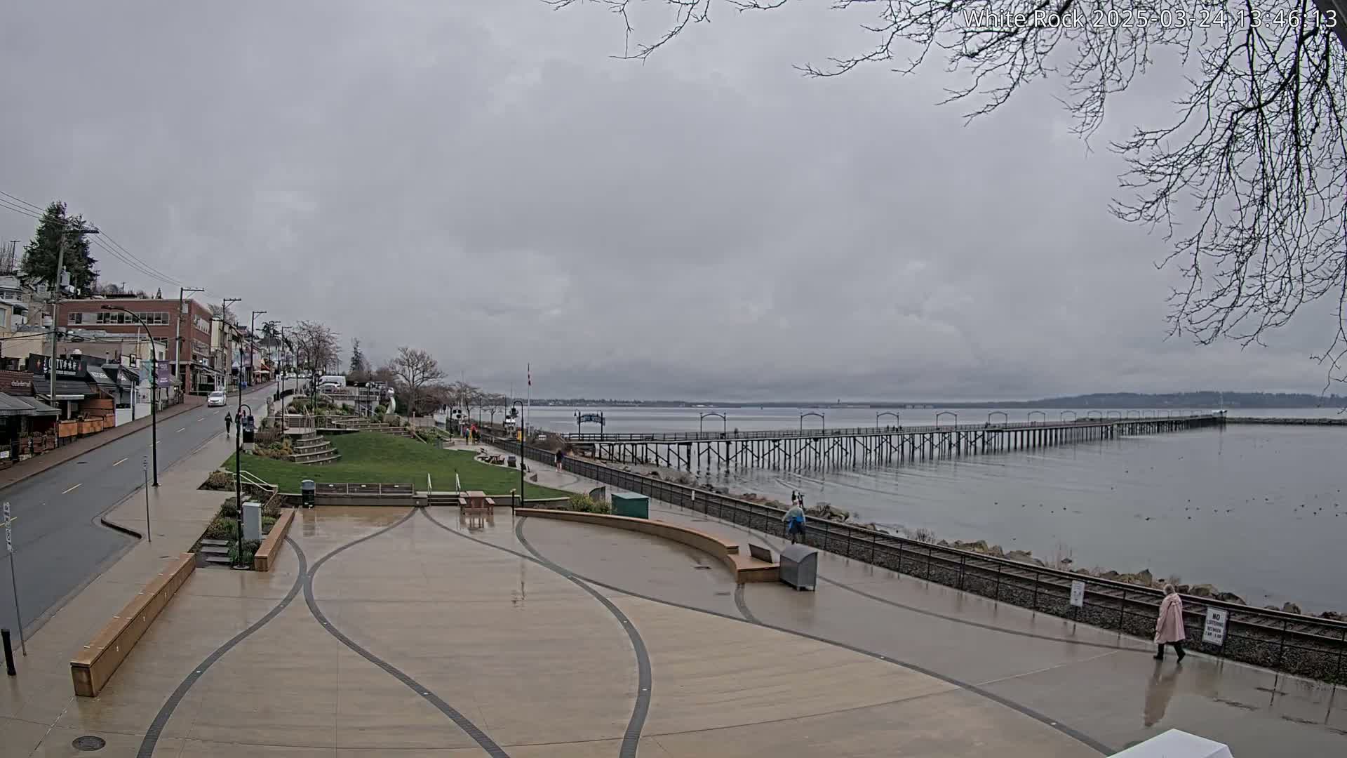 A long pier extends into a calm, gray body of water under an overcast sky, adjacent to a wet paved waterfront area with shops and a grassy park.