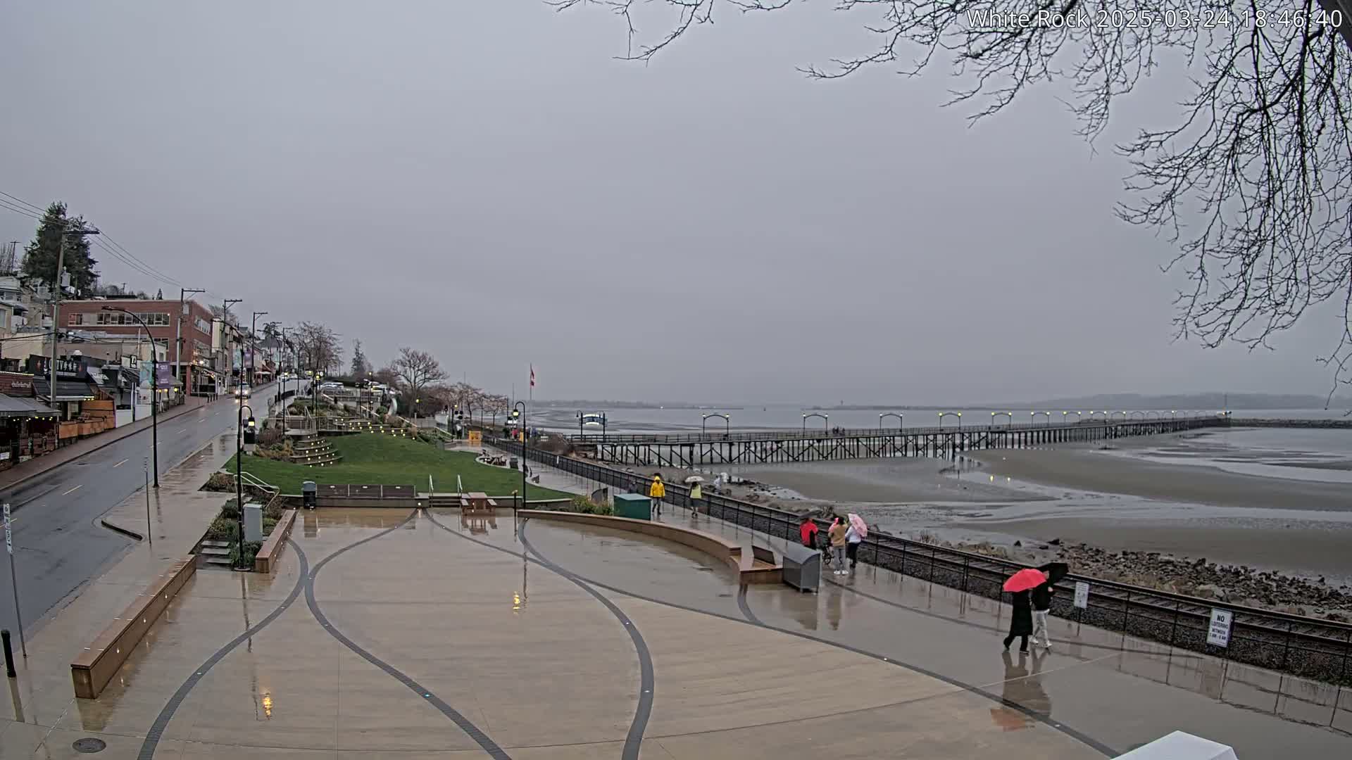 A wet, overcast day reveals a waterfront scene with a long pier extending into a calm bay, pedestrians strolling along a paved walkway near shops and a grassy area.