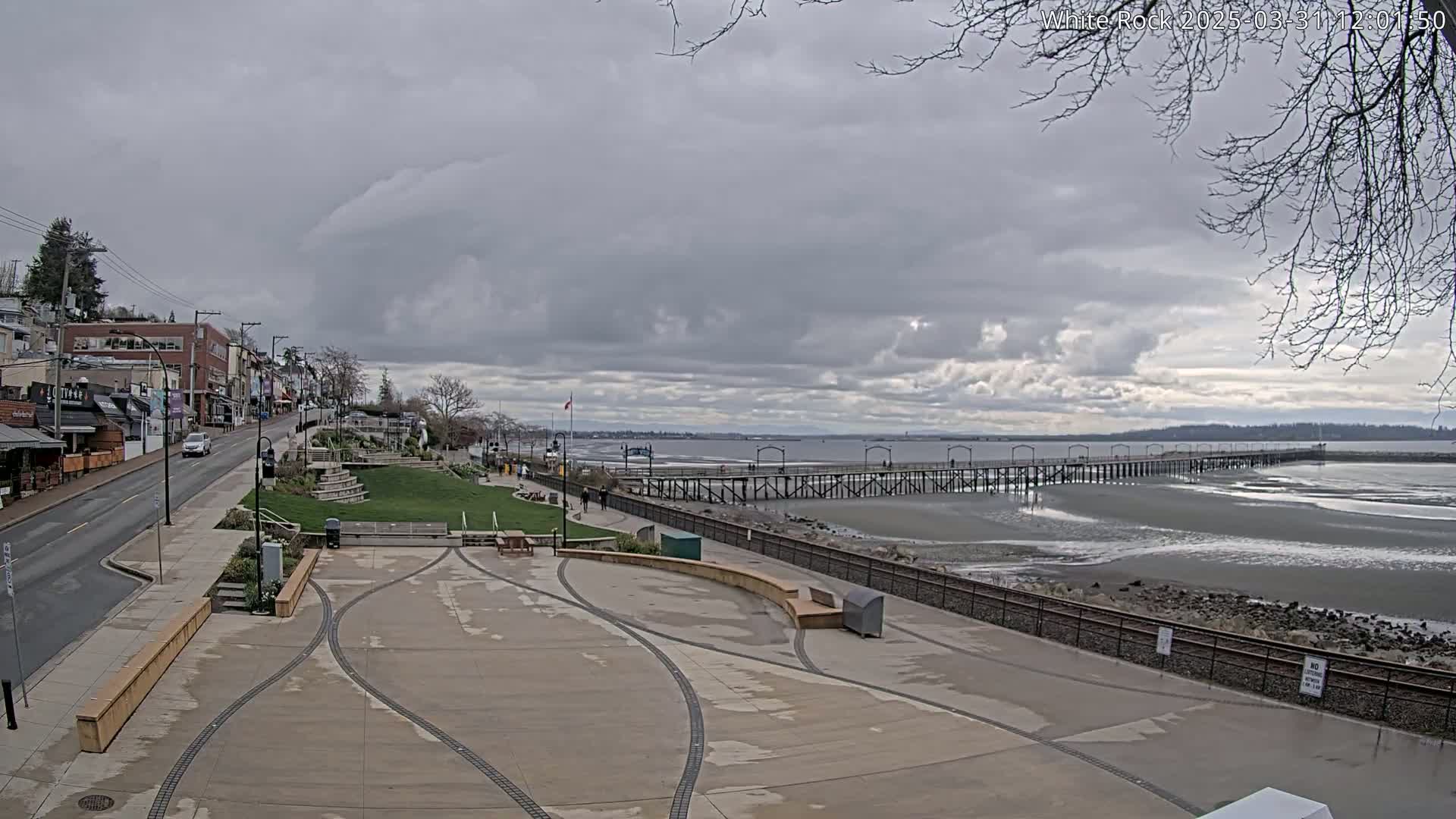 A long pier extends into a mostly calm, gray-toned body of water under a cloudy sky, adjacent to a paved waterfront area with a road and buildings behind it.
