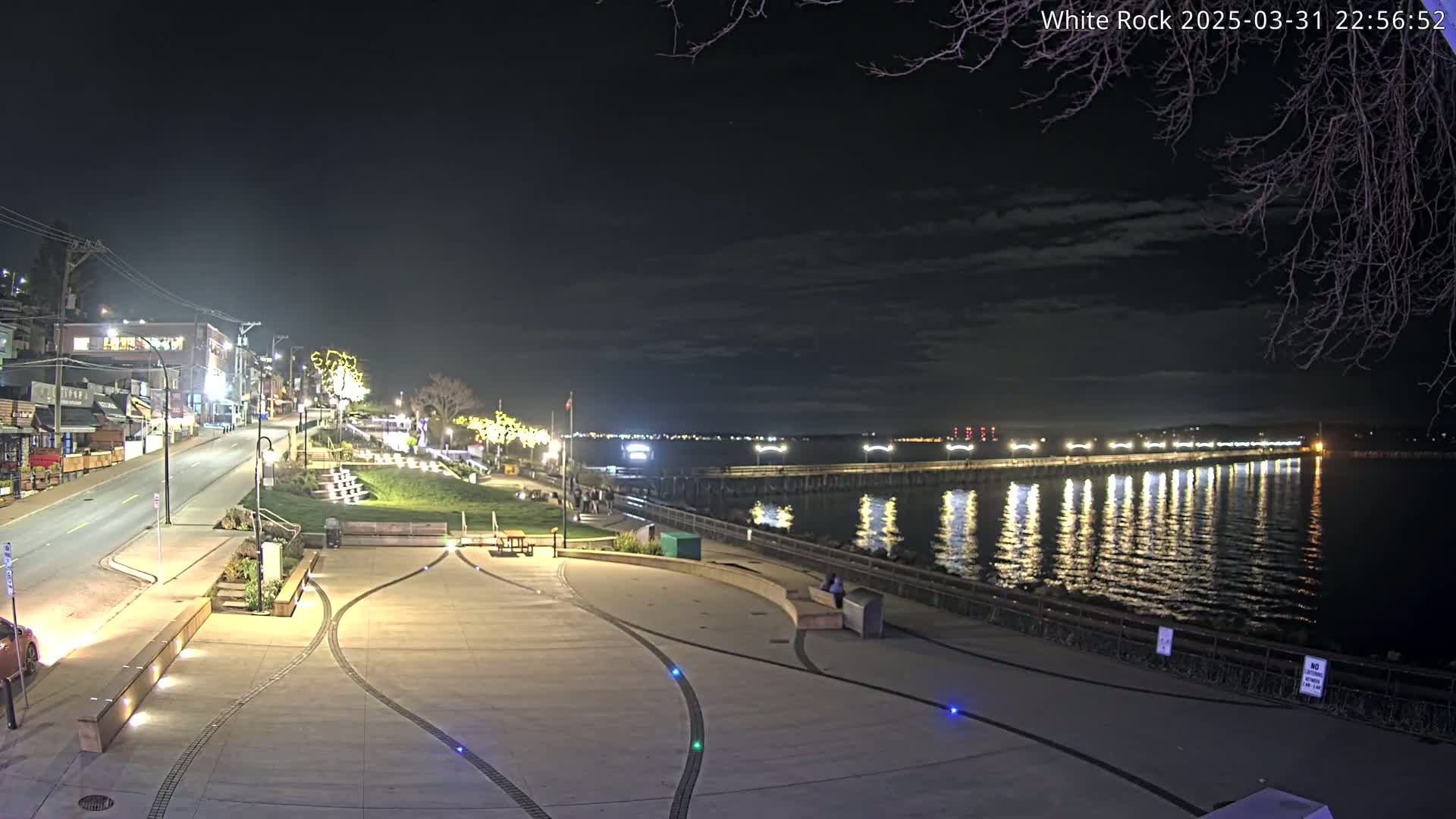 A nighttime view of a waterfront plaza with a long pier extending into calm water under a partly cloudy sky.