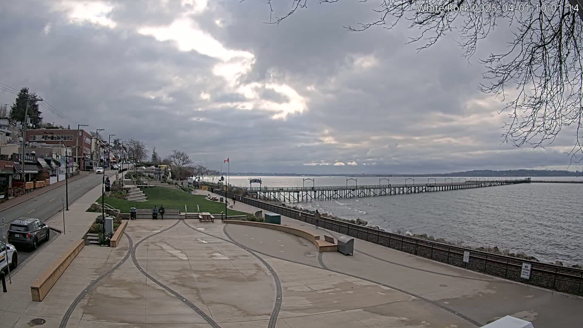 A long pier extends into a calm, gray-skied ocean, beside a paved waterfront area with shops and a grassy park in the foreground.
