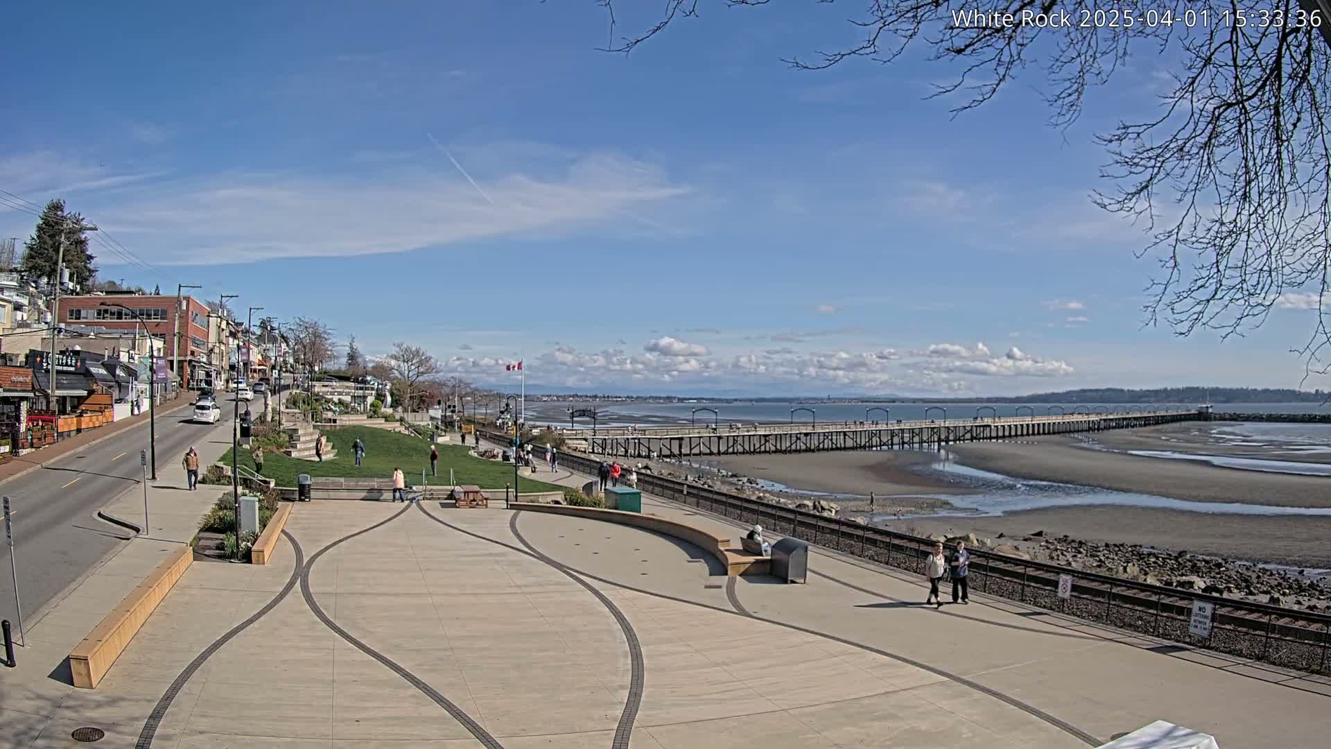 A sunny day reveals a seaside town with a long pier extending into a wide, sandy beach at low tide, and several people strolling along the paved waterfront walkway.