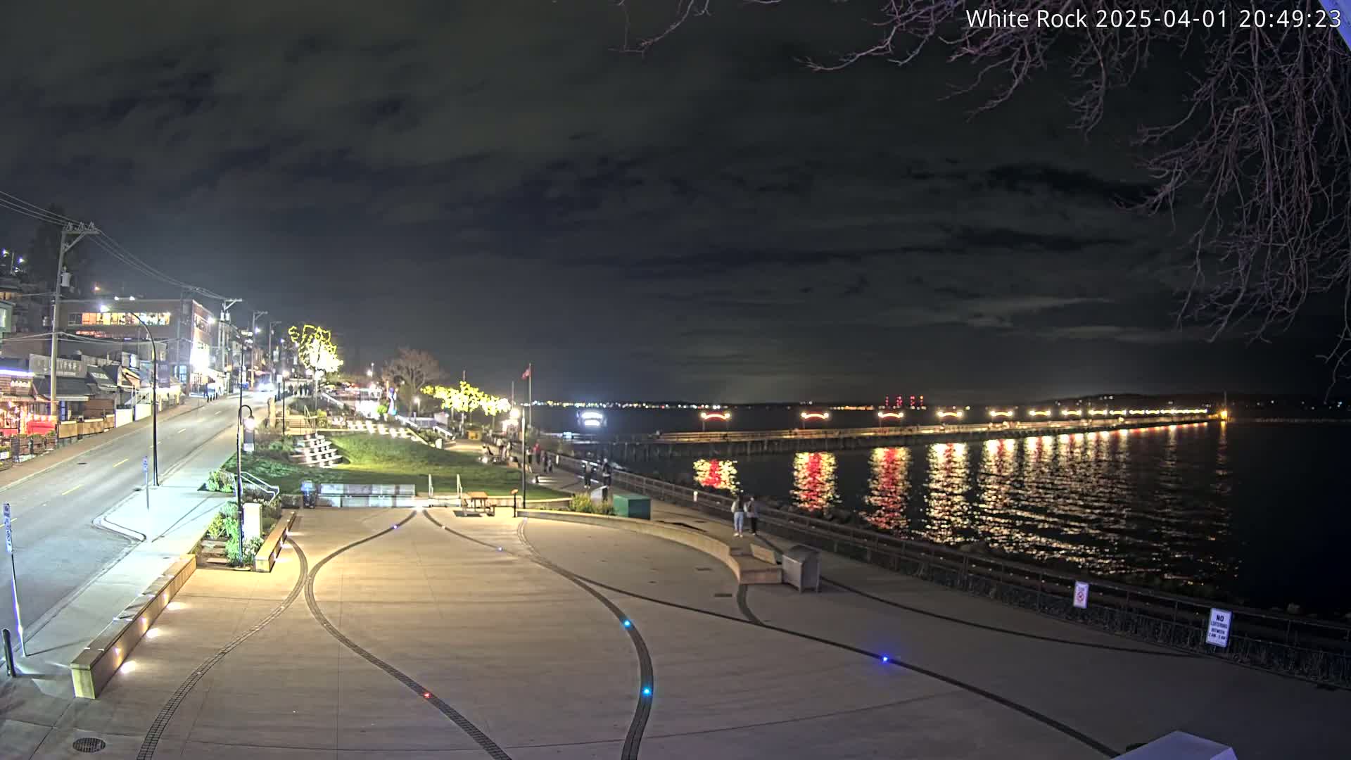 A nighttime view of a waterfront plaza with a street and buildings in the background, under a partly cloudy sky, shows a pier with lights extending into a calm body of water reflecting the lights.