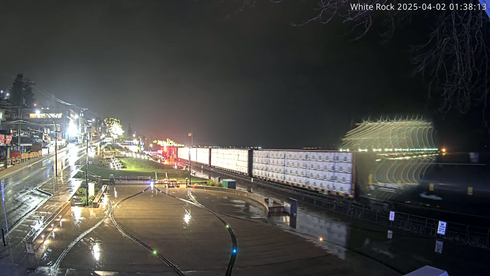 A long freight train passes by a rain-slicked waterfront town at night.