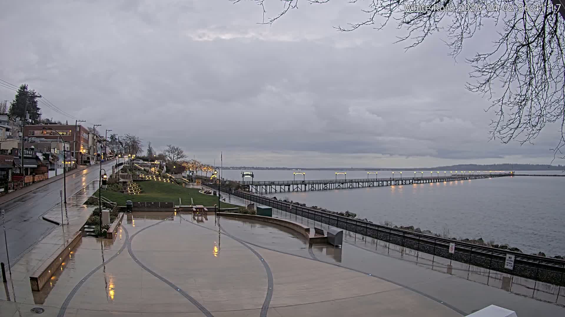 A long pier extends into a calm, gray body of water under an overcast sky, with a wet paved plaza and a street lined with buildings visible in the foreground.