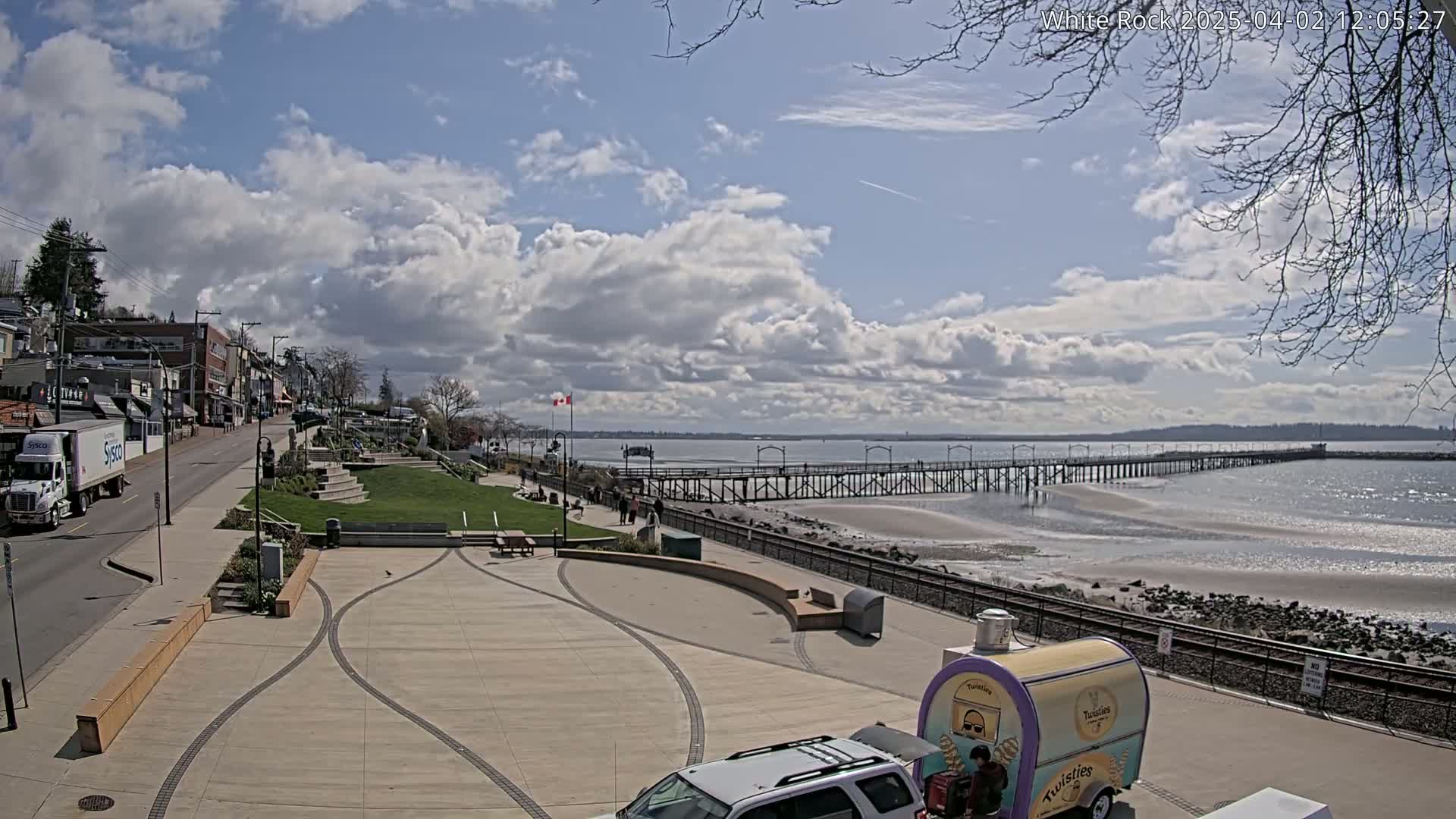 A seaside town with a long pier extends into a calm bay under a partly cloudy sky.