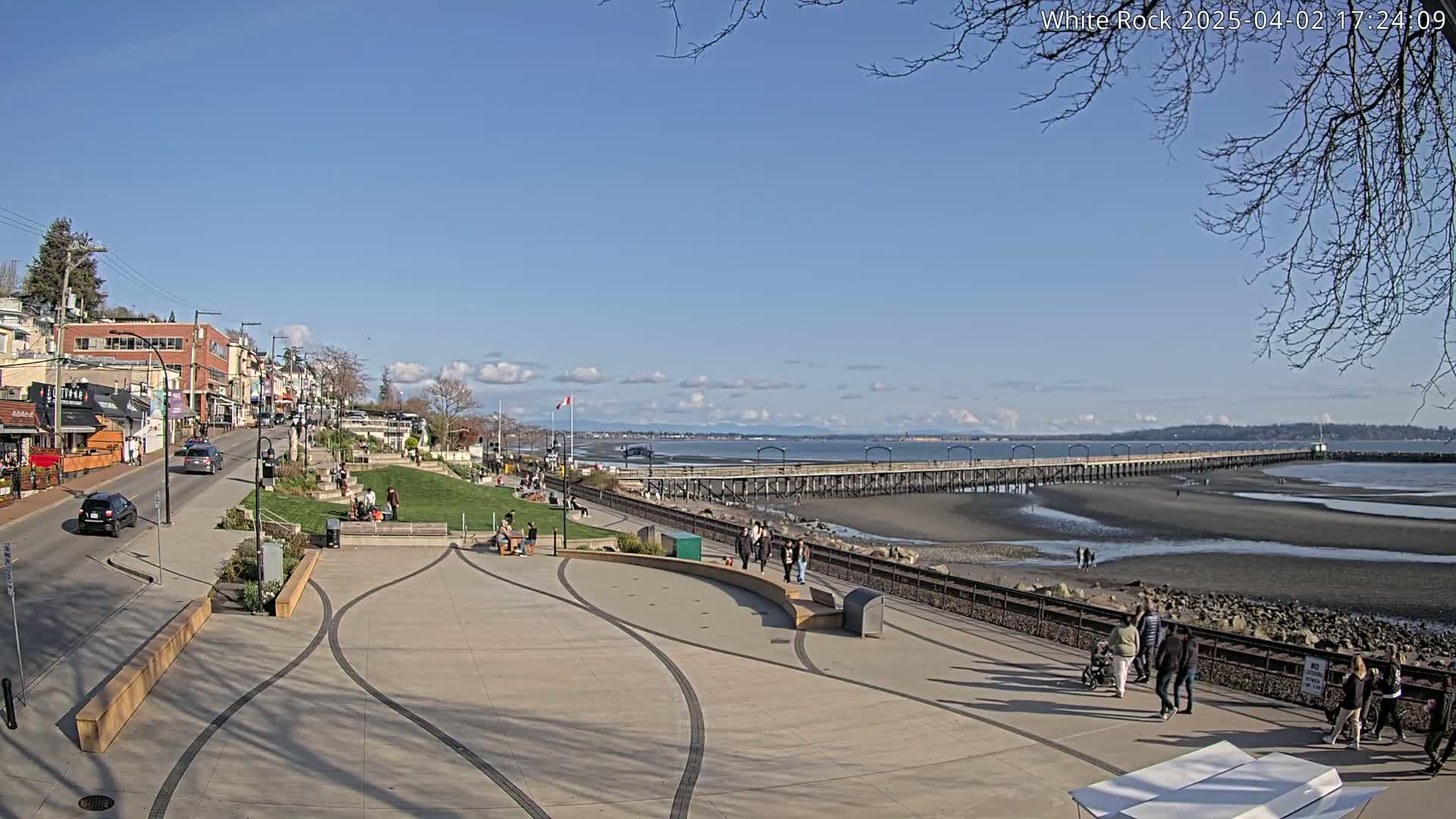 A sunny day reveals a seaside town with a long pier extending into a wide, sandy beach at low tide, people strolling along the promenade and sitting in a grassy park.
