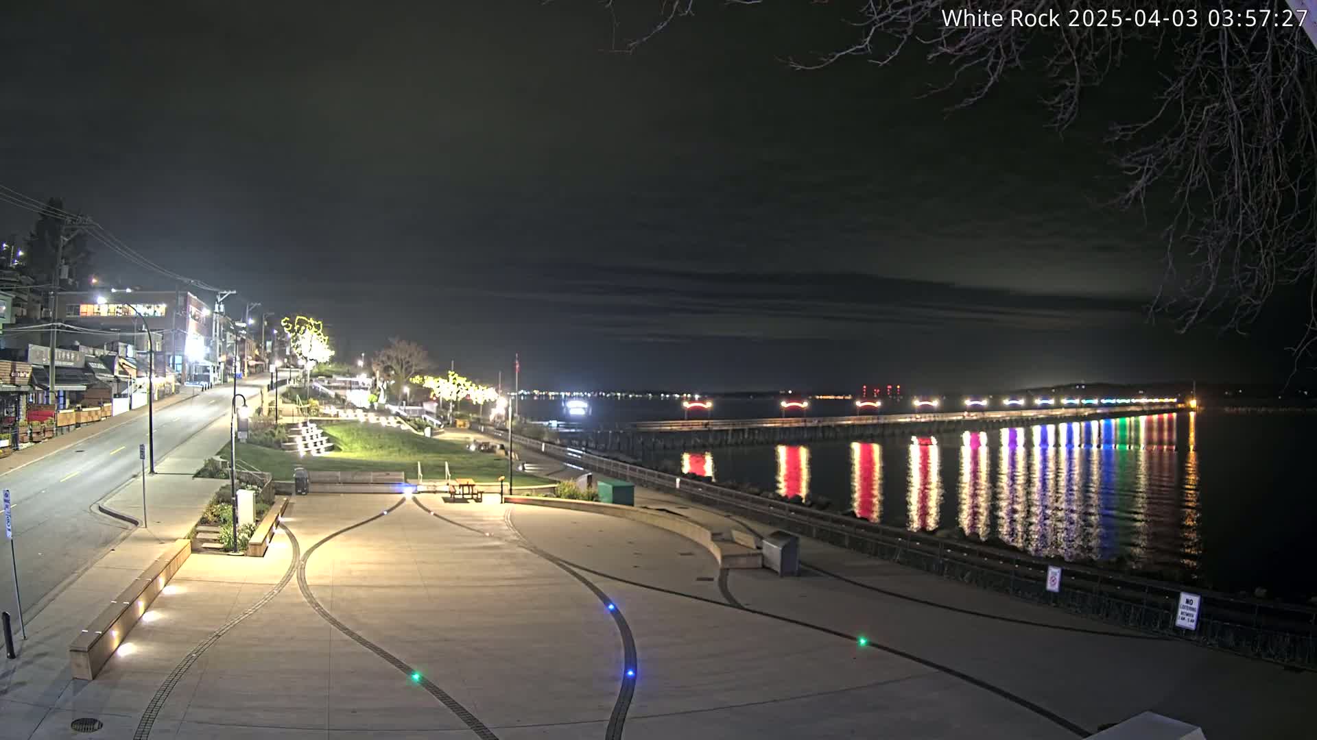 A brightly lit waterfront plaza at night overlooks a pier decorated with colorful lights reflected in the calm water.